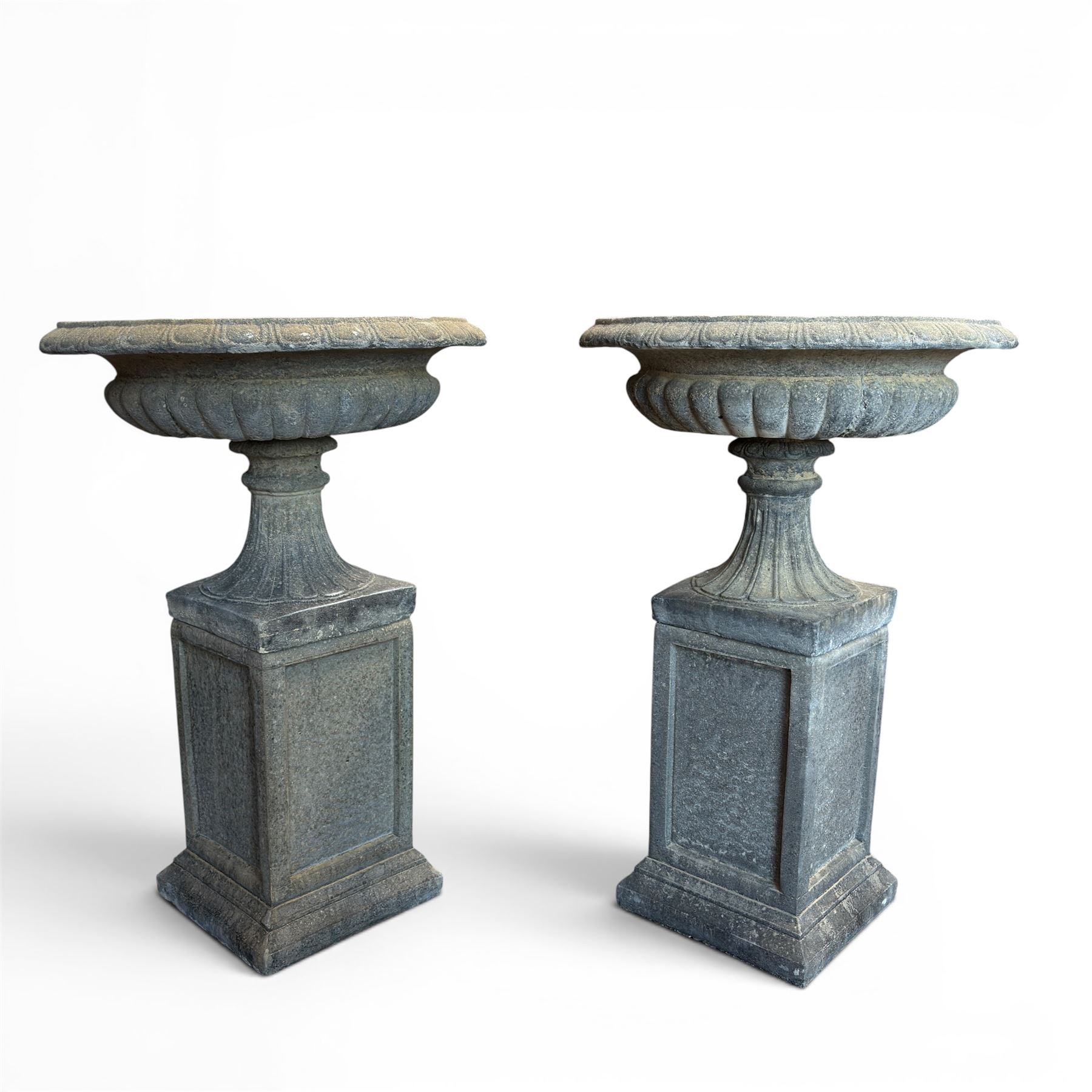 Pair of cast stone garden urns of kylix form on pedestals, the broad egg-and-dart moulded rim over a shallow gadrooned underbelly, with waisted tapering base, raised on matching plinth bases with faux panelled facia