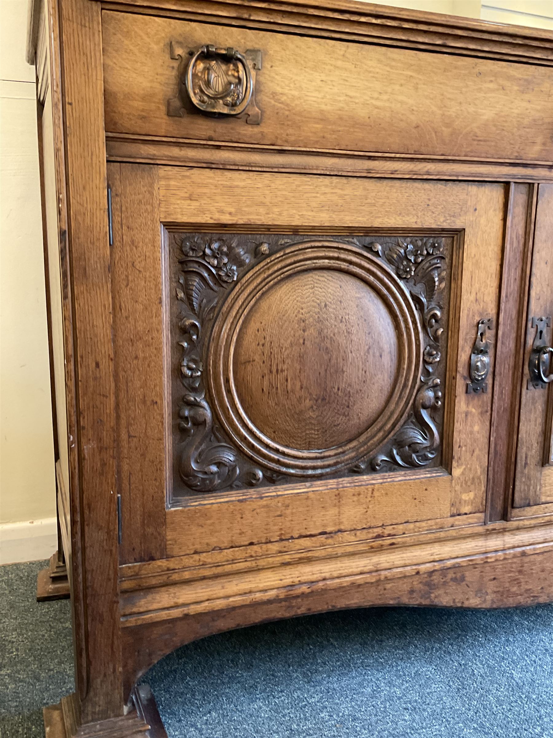 Late 19th century Arts & Crafts period oak mirror back sideboard, shaped bevelled mirror back carved with foliate scrolls and flower heads, down swept supports with elongated club-shaped uprights, moulded rectangular top over two drawers and three cupboards, the drawer fronts with copper handles decorated with sailing ships, panelled doors carved with mythical beasts, flowerheads and foliage, on moulded block feet