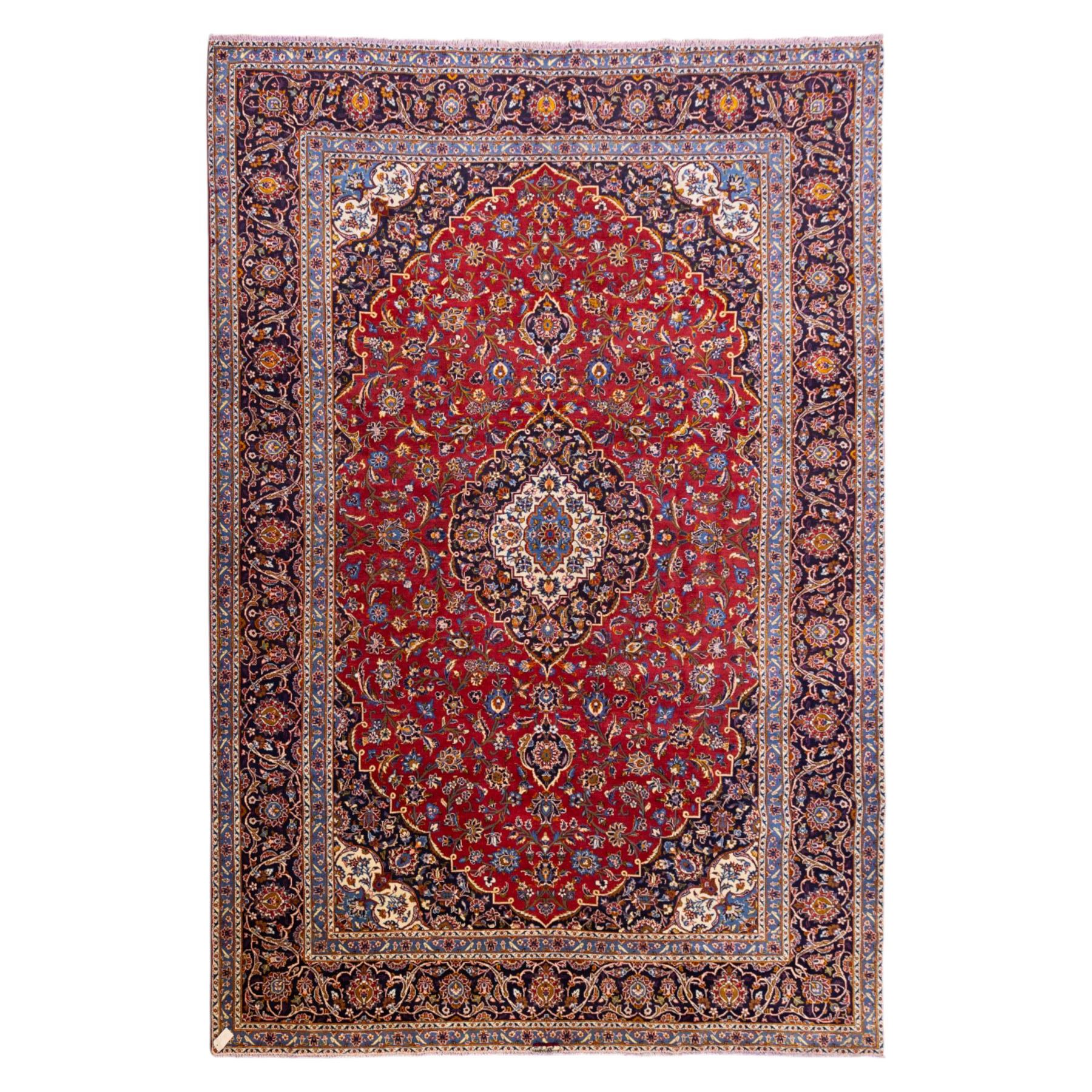 Central Persian Kashan hand-knotted carpet, rich crimson red field with a central cusped medallion with finely drawn floral sprays surrounded by dense scrolling vine work and palmettes, framed by a broad indigo ground border of flowering arabesques flanked by multiple guard stripes, in a palette of red, blue, ivory, sage green, and soft ochre tones, with cotton fringe ends    