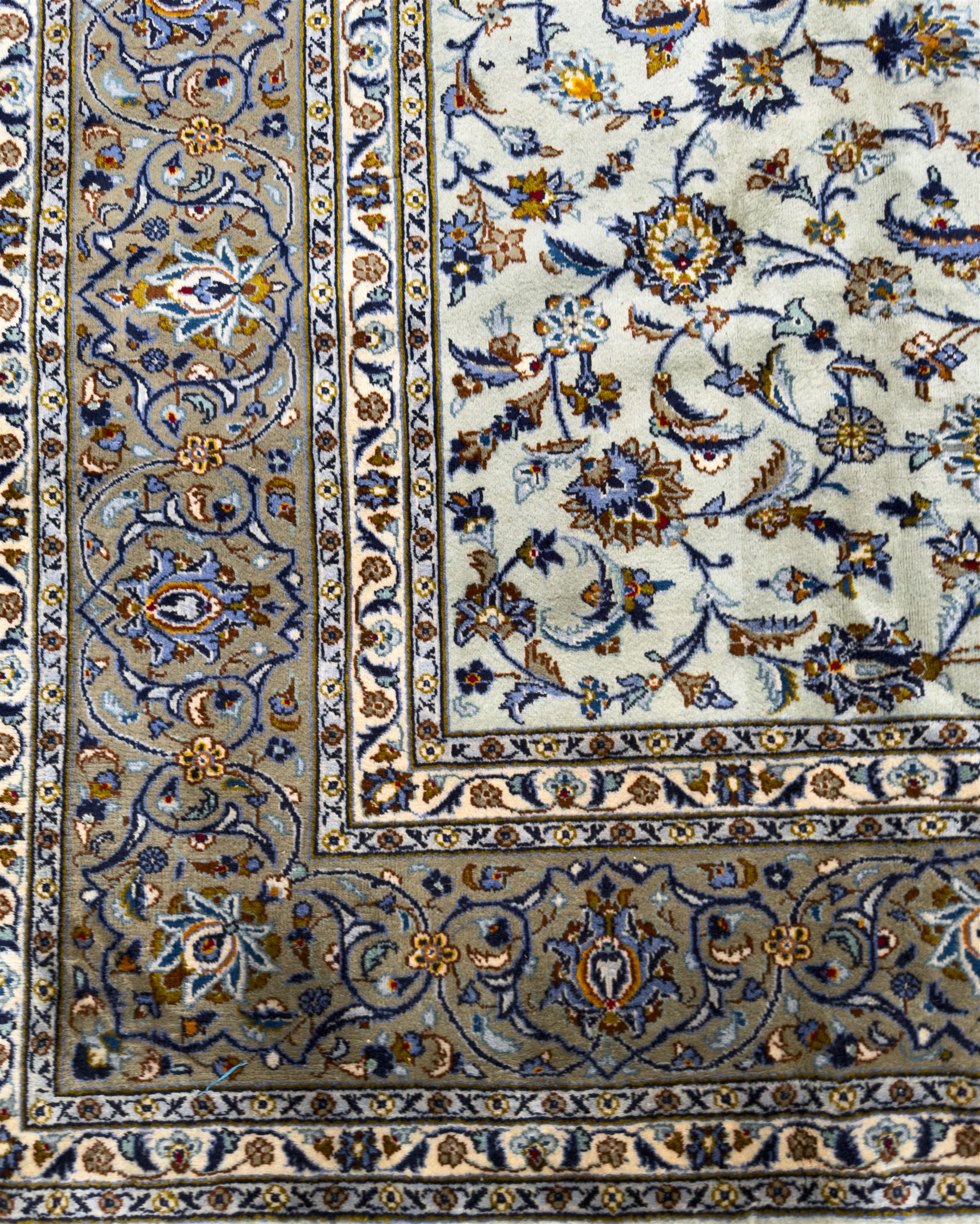 Central Persian Kashan hand-knotted carpet, the ivory ground intricately decorated with scrolling vines, palmettes, and stylised flowerheads in soft blue, gold and brown tones, enclosed by a broad indigo border with repeating floral motifs and multiple guard stripes