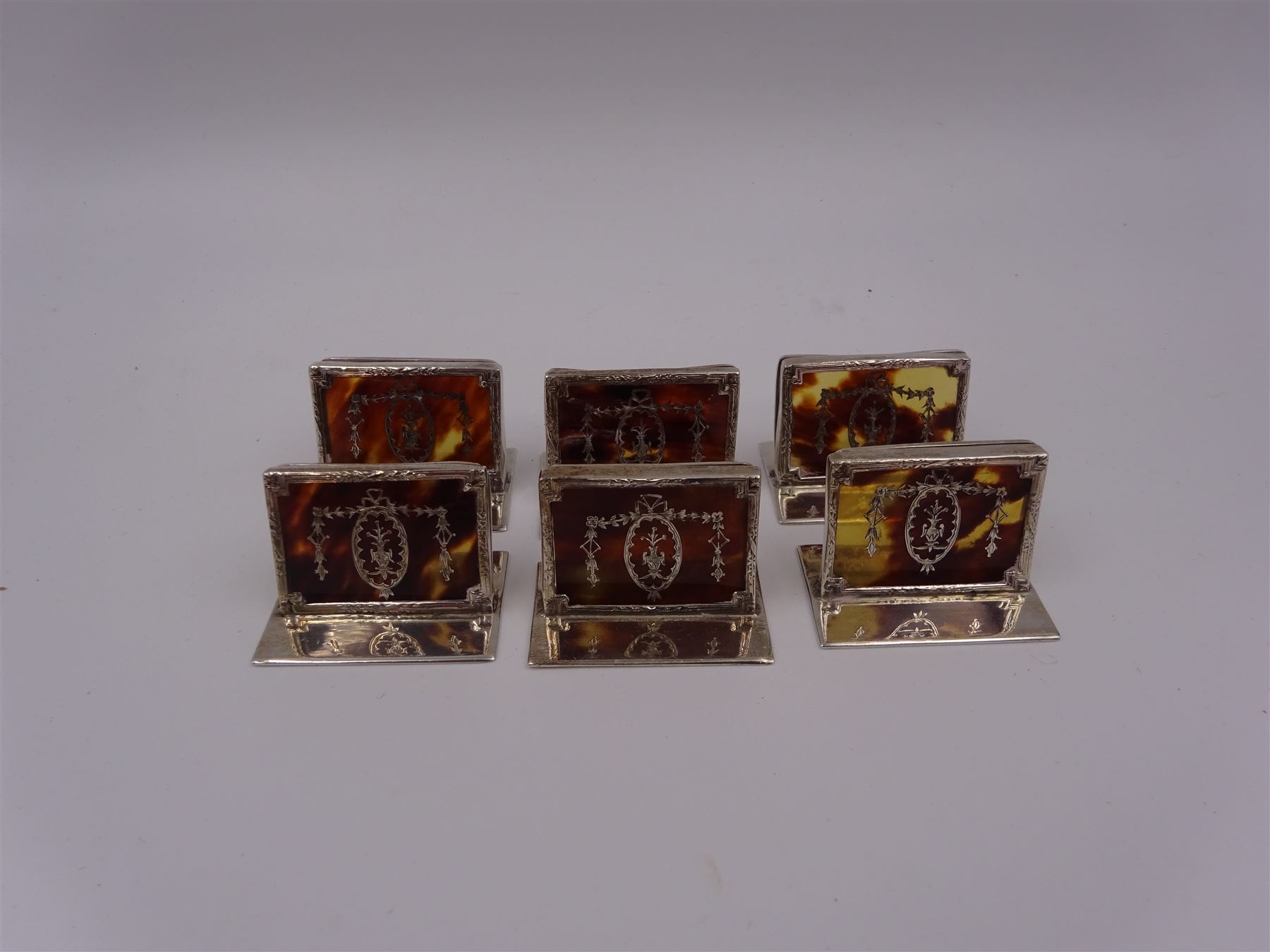 Set of six early 20th century silver mounted tortoiseshell place card holders by Asprey, the rectangular silver reed and ribbon frame bordering a central tortoiseshell panel inlaid with a silver floral motif, upon an oblong foot, hallmarked Asprey & Co Ltd, London 1913, H3cm, contained within a tooled leather fitted case with velvet and silk lined interior 