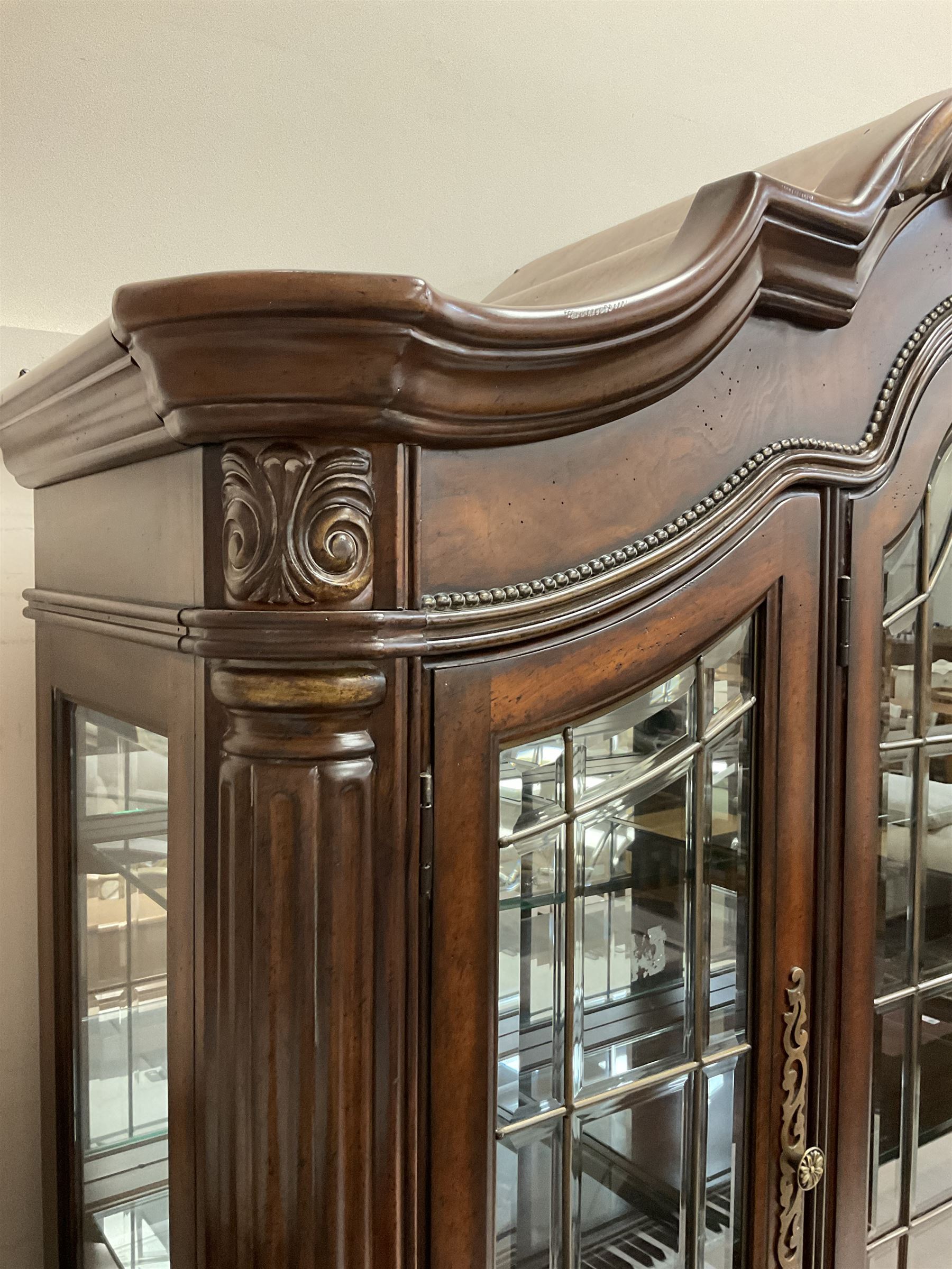 Kevin Charles - American walnut display cabinet, illuminated interior, shell carved pediment above four bevel edge doors enclosing six glazed shelves above one central long drawer flanked by two short drawers, above three cupboards, carved bracket supports 