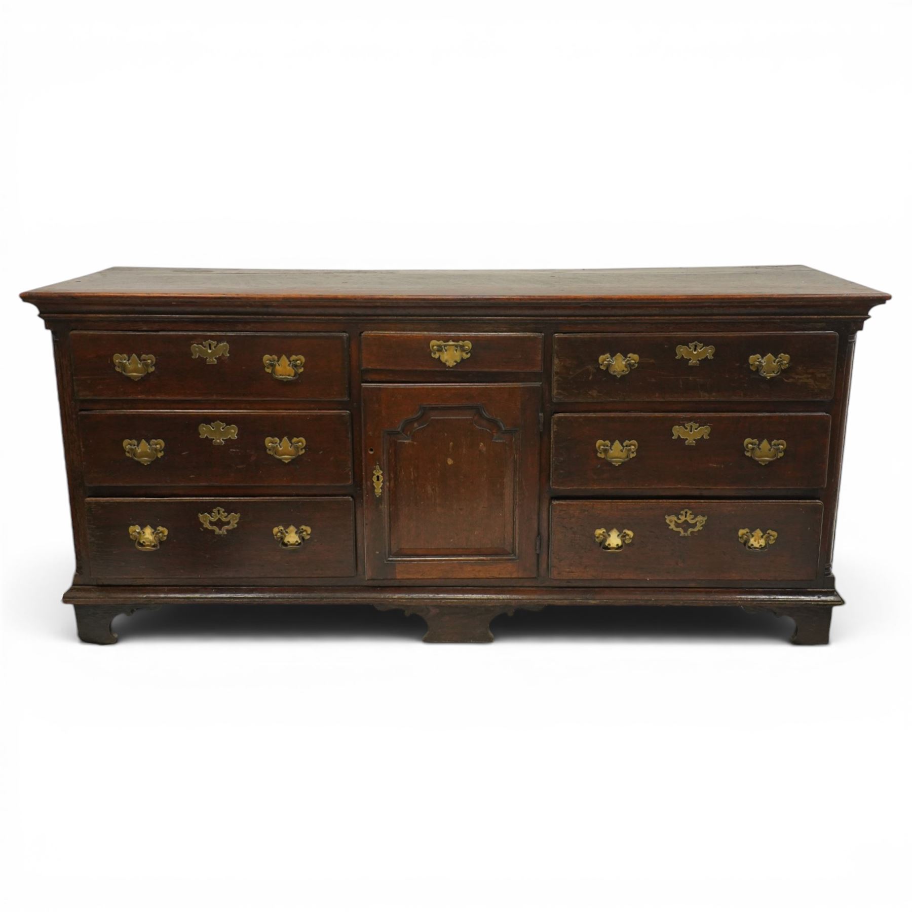 19th century Georgian oak long dresser base, rectangular plank top above a central frieze drawer over a fielded panel cupboard, flanked by three banks with quarter cantered pilasters, brass swan neck handles and escutcheons, raised on shaped bracket feet