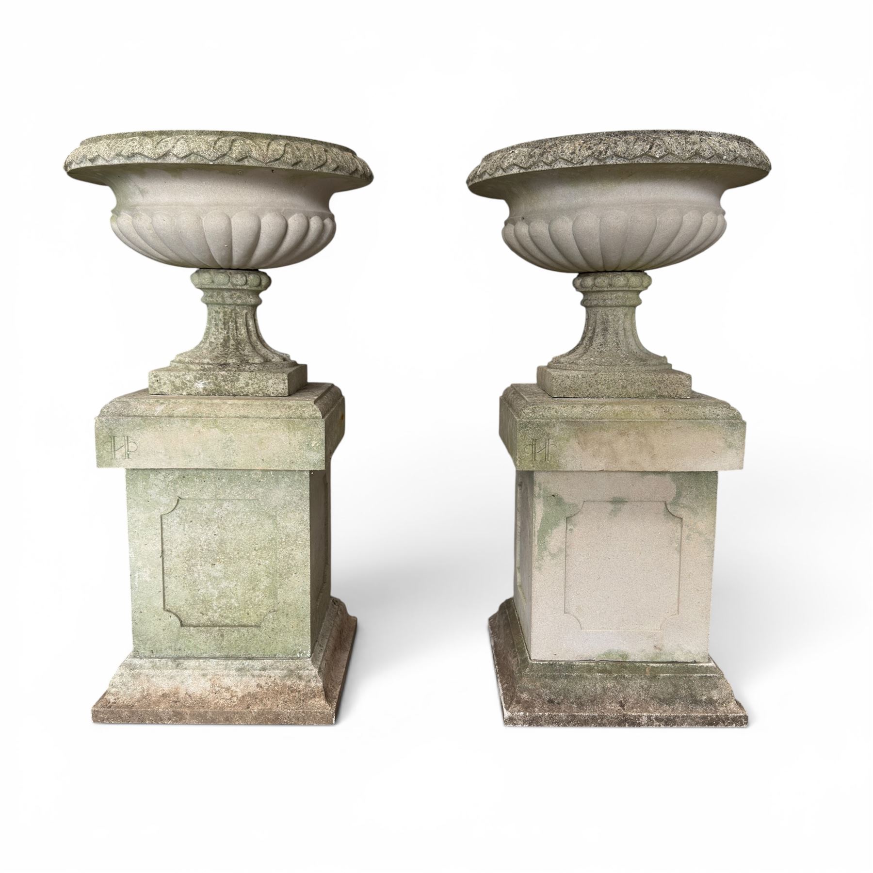 Pair of weathered cast stone Neoclassical design garden Kylix shape urns, foliate moulded rim over a gadrooned underbelly, the scalloped socle over a rectangular pedestal with faux-panelled sides, stamped with stylised ‘H’ monogram