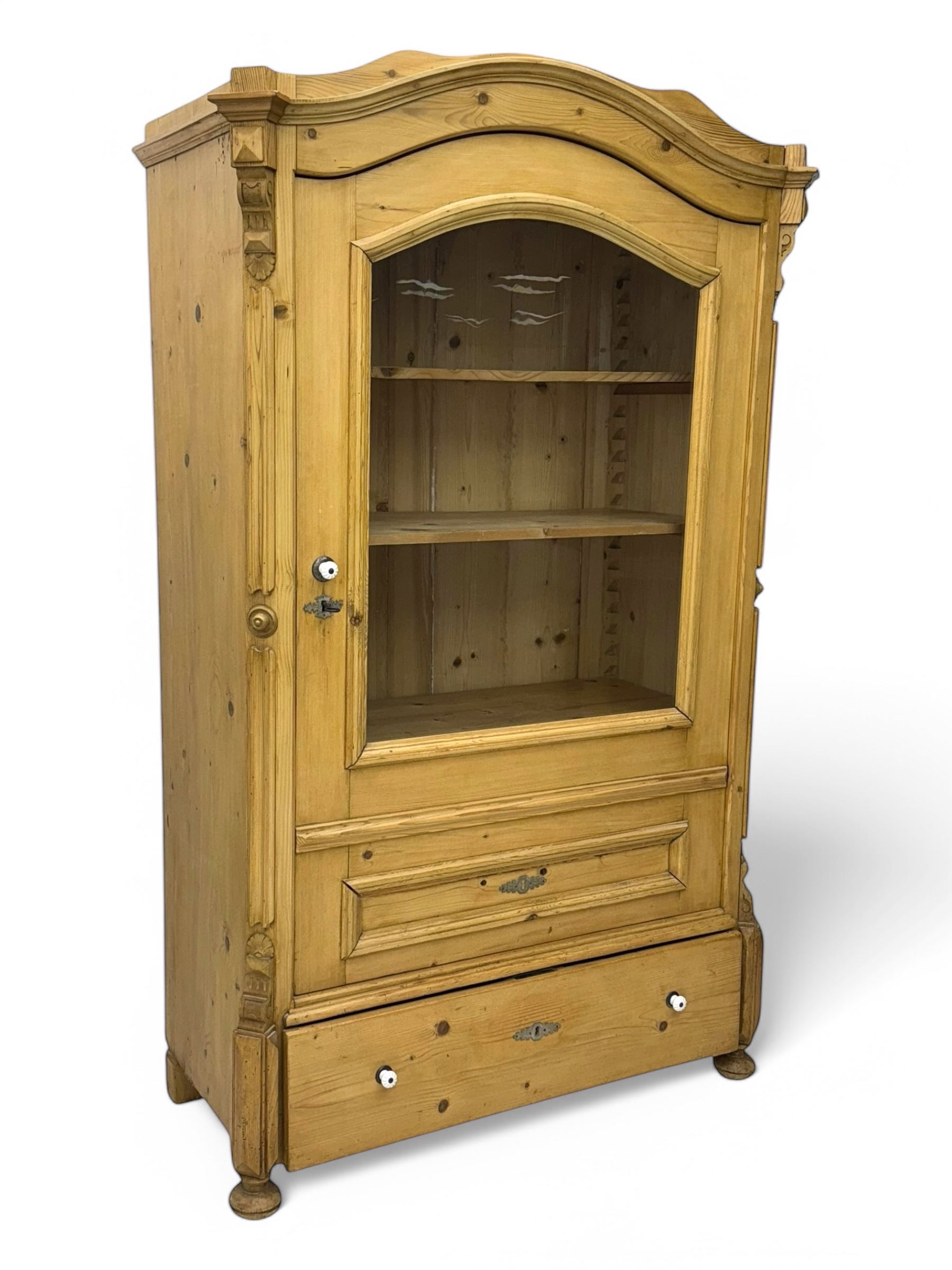 Late 19th century Continental pine vitrine, shaped and moulded arched cornice over a single glazed door enclosing adjustable shelves, flanked by carved pilasters with foliate and geometric motifs, fitted with single moulded drawer, raised on compressed bun feet