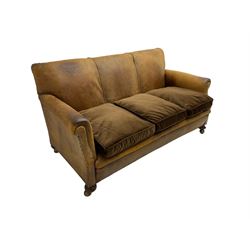 Early-to-mid-20th century three seat sofa, upholstered in worn tan leather with studwork, loose cushions upholstered in brown fabric, on compressed bun feet with castors
