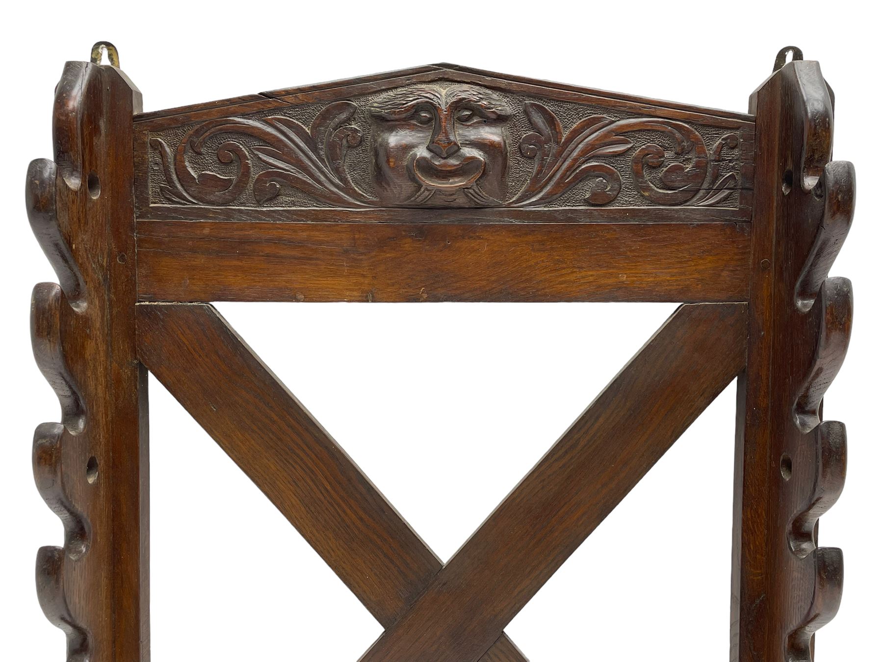 Victorian oak wall hanging whip or stick rack, sloped arch pediment carved with central grotesque mask with extended scrolling foliage, the upright supports cut with rests, x-framed back, two lower hooks