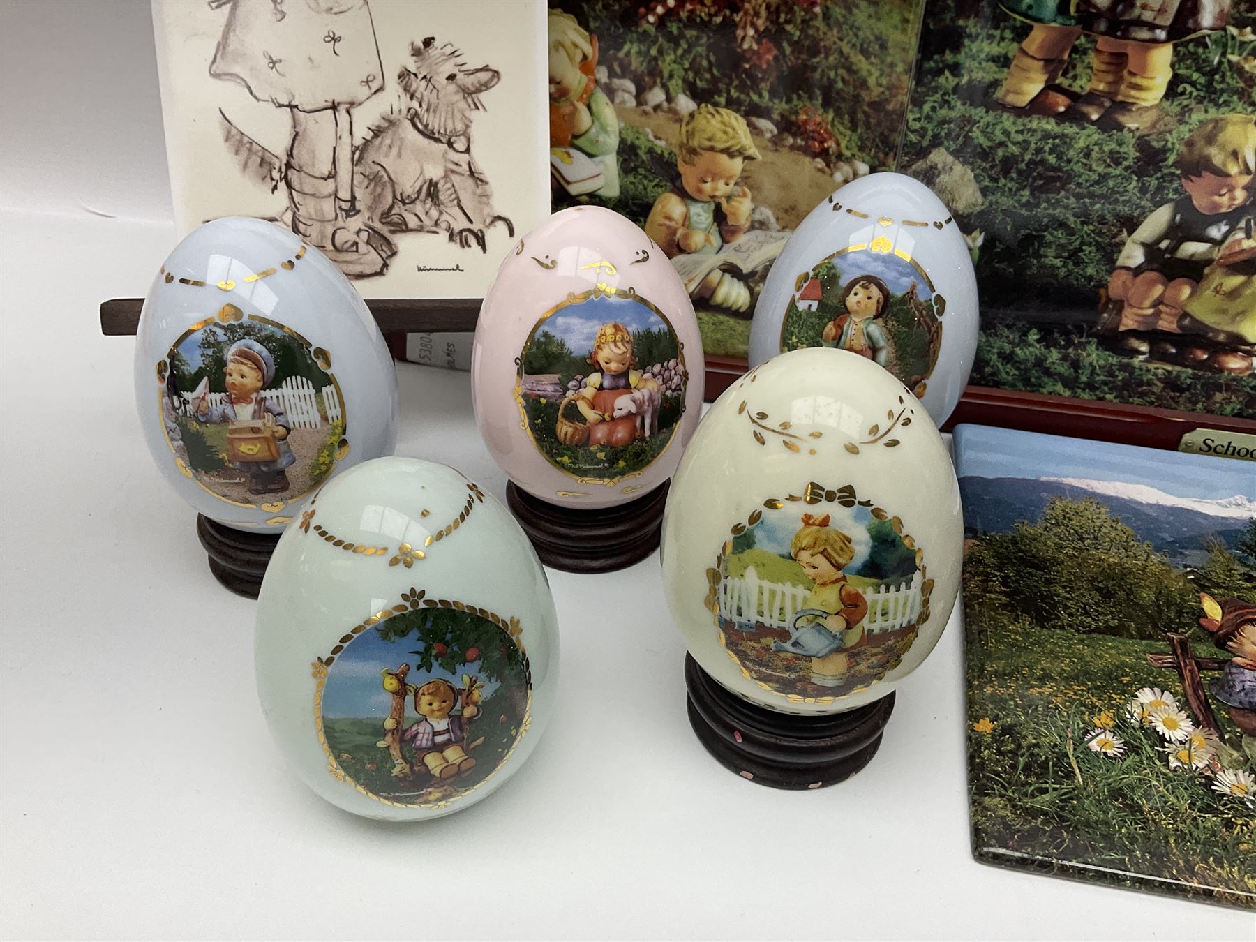 Two ceramic Hummel postcards by Goebel, both displayed on wooden easels, together nine Hummel eggs, set of four Hummel School Days plates, on wooden hanging rack, Goebel Collectors Club Member plaque etc