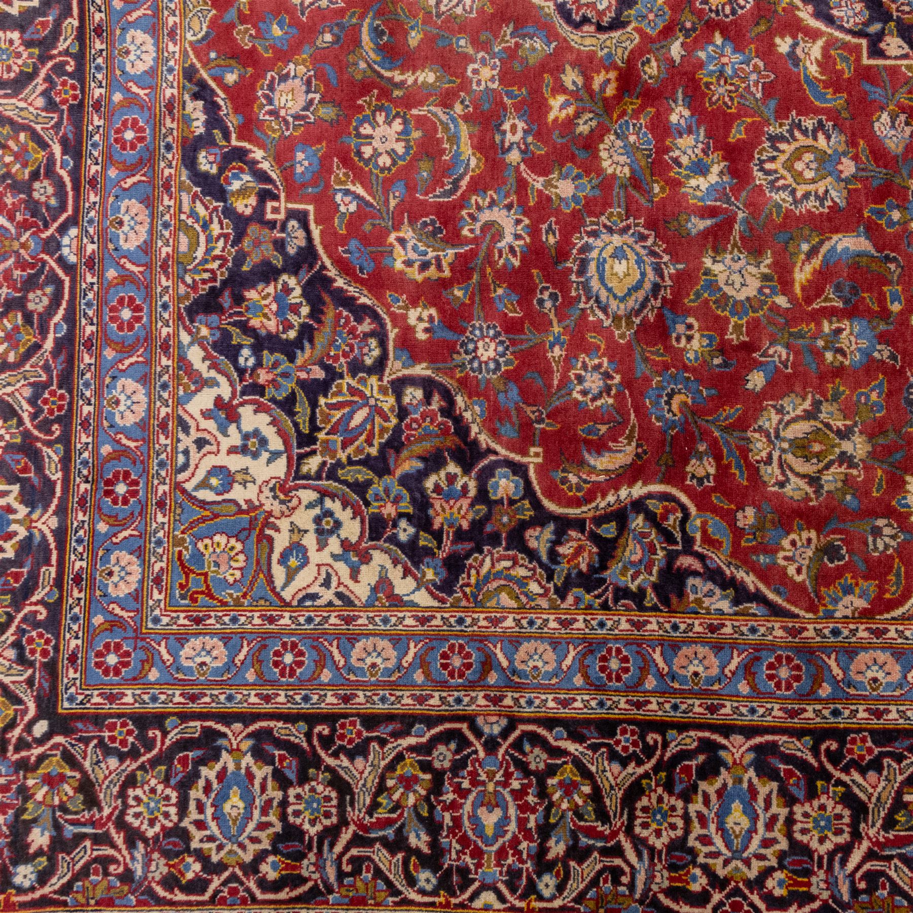 Central Persian Kashan hand-knotted carpet, madder red field with a central cusped medallion of ivory and deep indigo ground, enclosing a stylized floral rosette, framed by scrolling arabesques, field richly ornamented with an intricate network of flowering vinery, palmettes, and delicate floral sprays, spandrels in deep indigo, principal border of navy ground decorated with a continuous palmette and vine-scroll, flanked by multiple guard stripes, with cotton fringe ends 