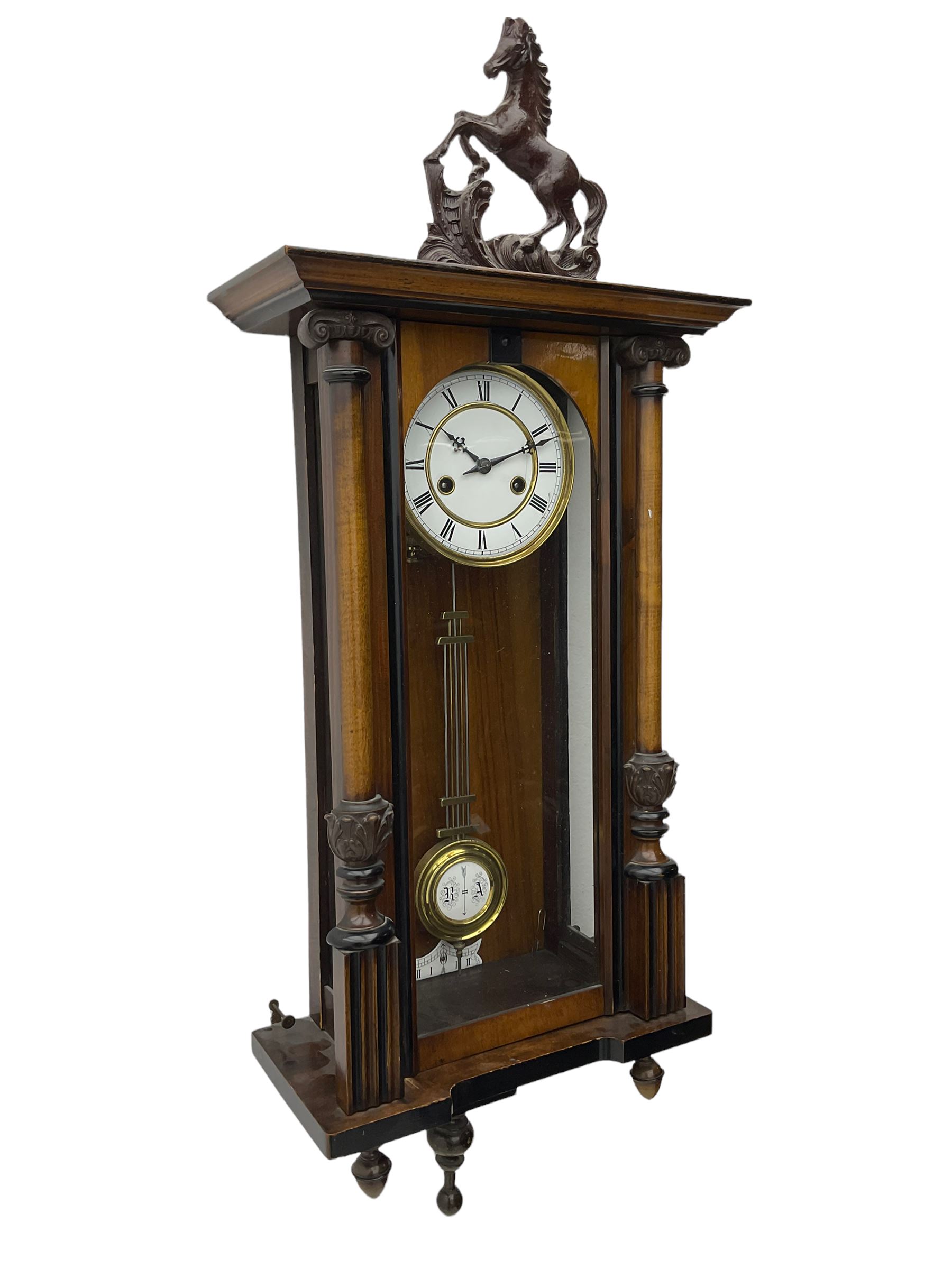 A late 19th century German spring driven 'Vienna' wall clock with a fully glazed door flanked by turned columns with ionic capitals and carving, replacement pediment depicting a prancing horse, with a two-piece enamel dial, Roman numerals, minute track and gothic steel hands, 8-day movement striking the hours and half hours on a gong,  with a gridiron pendulum and beat plate. With key.


