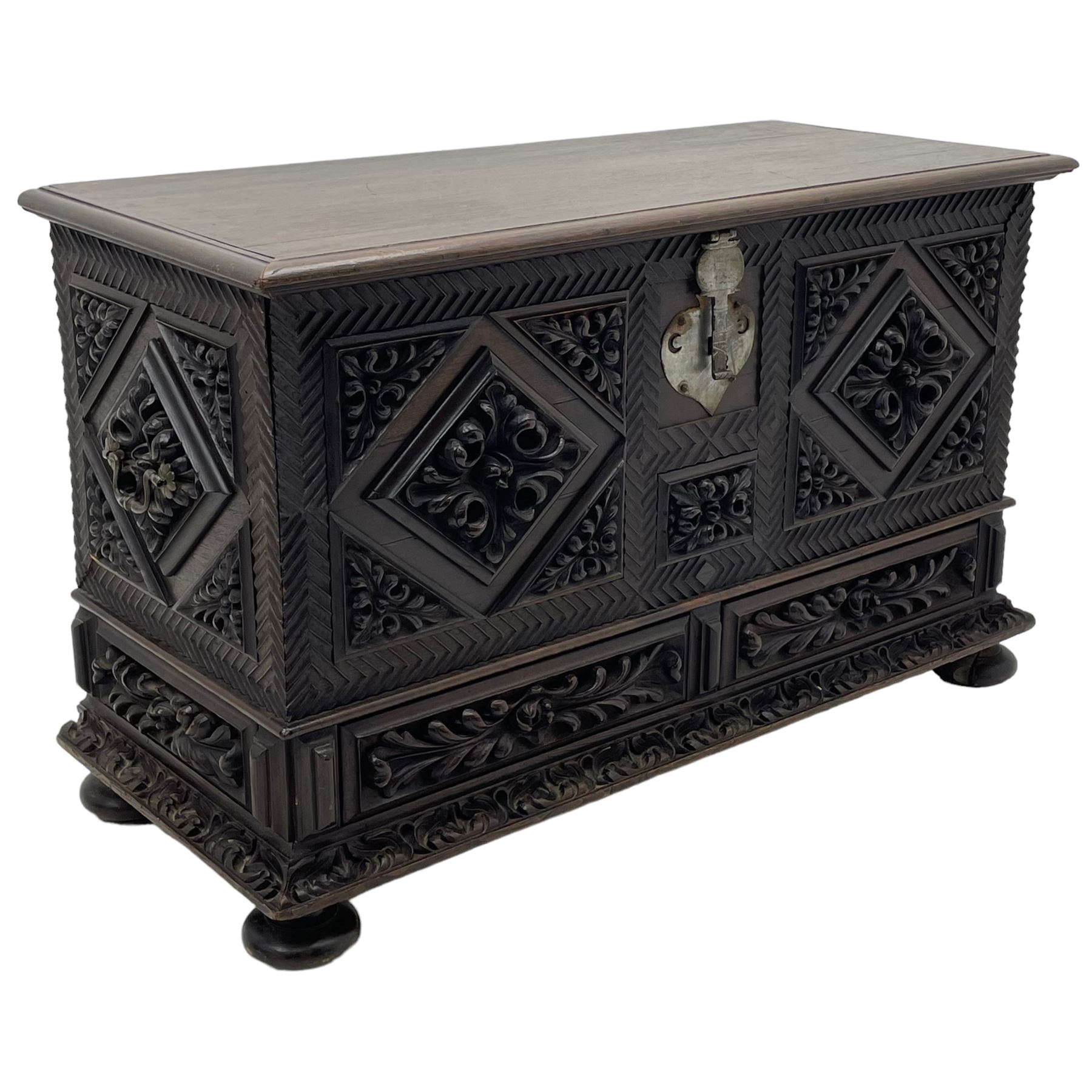Early 20th century carved hardwood kist or mule chest of small proportions, moulded rectangular hinged lid over foliage carved geometric lozenge panels, the frame carved with chevron banding, fitted with two drawers, the drawer fronts carved with extending leaf motifs over a foliage carved lower moulding, on turned feet