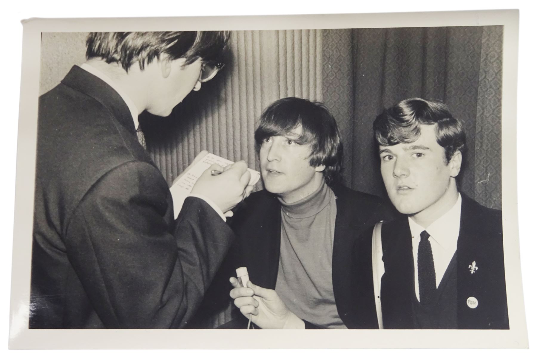 John Lennon interview recording conducted by John Hill student of Hull University on the 16th October 1964 at the A.B.C. Theatre Hull on behalf of the university newspaper 'Torchlight'. The interview lasts eight and a half minutes. It catches John in an upbeat breezy mood answering questions about the following topics: the song writing process ('sometimes you get an idea ....I dunno....my left leg.... and then write about that....'), recording (.... 'they just say - you're recording on such and such a date and they hope we turn up with some good songs written ....'), an alternative career path (.... 'I'd probably just have been a lay-about....'), failing at college (....'some other lads did my exams for me ....'), reading matter ( Clockwork Orange, Alice in Wonderland), his books of prose, tastes in music, the Blues, The Rolling Stones, being a Beatle vis-a-vis anonymity ('Whatever I'd be I'd choose to be rich .... who wants to walk down the street?').
The interview was recorded on 1/4 inch Emitape on a Fi-Cord Fidelity Recording machine. The reel-to-reel tape comes in its original Emitape plastic box and is accompanied by the Fi-Cord reel-to-reel machine on which the interview was originally recorded. Also included in this lot are a copy of the 'Torchlight' newspaper from 21st November 1964, two photographs of the interviewer with John Lennon (the interviewer is pictured to the left of John Lennon in the photograph in which John is signing a sketch) and one photograph of Paul McCartney with another reporter, the largest measures 12 x 16.5cm and a transcript of the interview. The recording is also accompanied by the original Fi-Cord tape recorder with microphone, four original 2v batteries and battery charger. The tape recorder has been adapted to take 3xAA batteries and the 2v battery charger is fitted with a standard UK 3-pin AC plug. The interview was recorded in the press room prior to the Beatles concert at the A.B.C. Theatre and is sold with copyright.