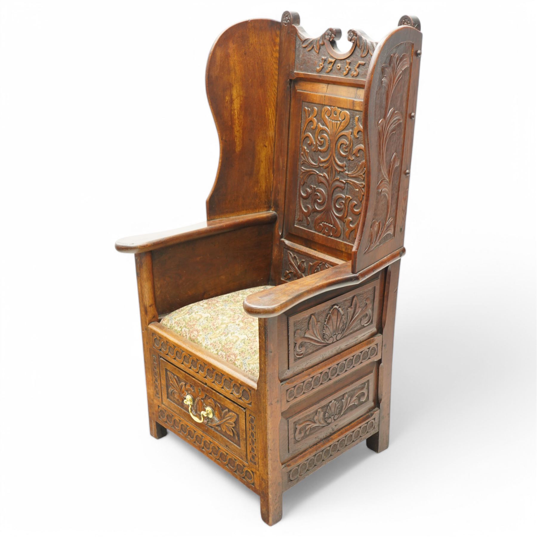 18th century design oak lambing chair, flower head and foliate carved cresting inscribed '1715' over panelled back carved with scrolling leafy branches, enclosed by shaped wingbacks over arms and panelled sides, the side panels carved with shells and scrolled leafage, upholstered seat over single drawer with matching carved decoration, on stile supports 
