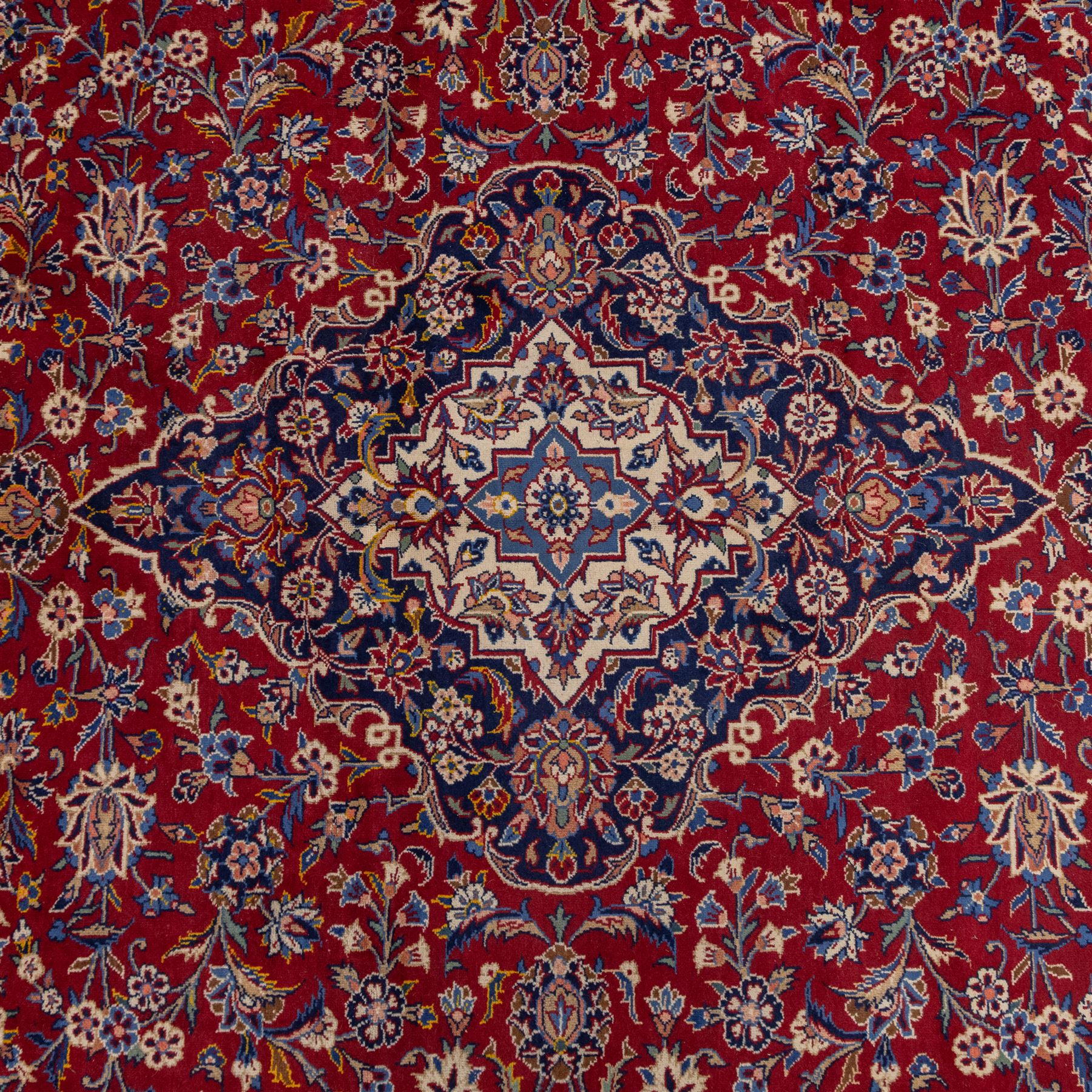 Central Persian Kashan hand-knotted carpet, madder red field with central cusped medallion in navy and ivory enclosing a floral rosette and radiating scrolling arabesques and palmettes, across a densely ornamented ground of finely drawn vinery enriched with a profusion of flowering sprays and stylized blossoms, spandrels in deep indigo and principal border of navy ground decorated with a continuous palmette and vine-scroll flanked by multiple guard stripes, with cotton fringe ends