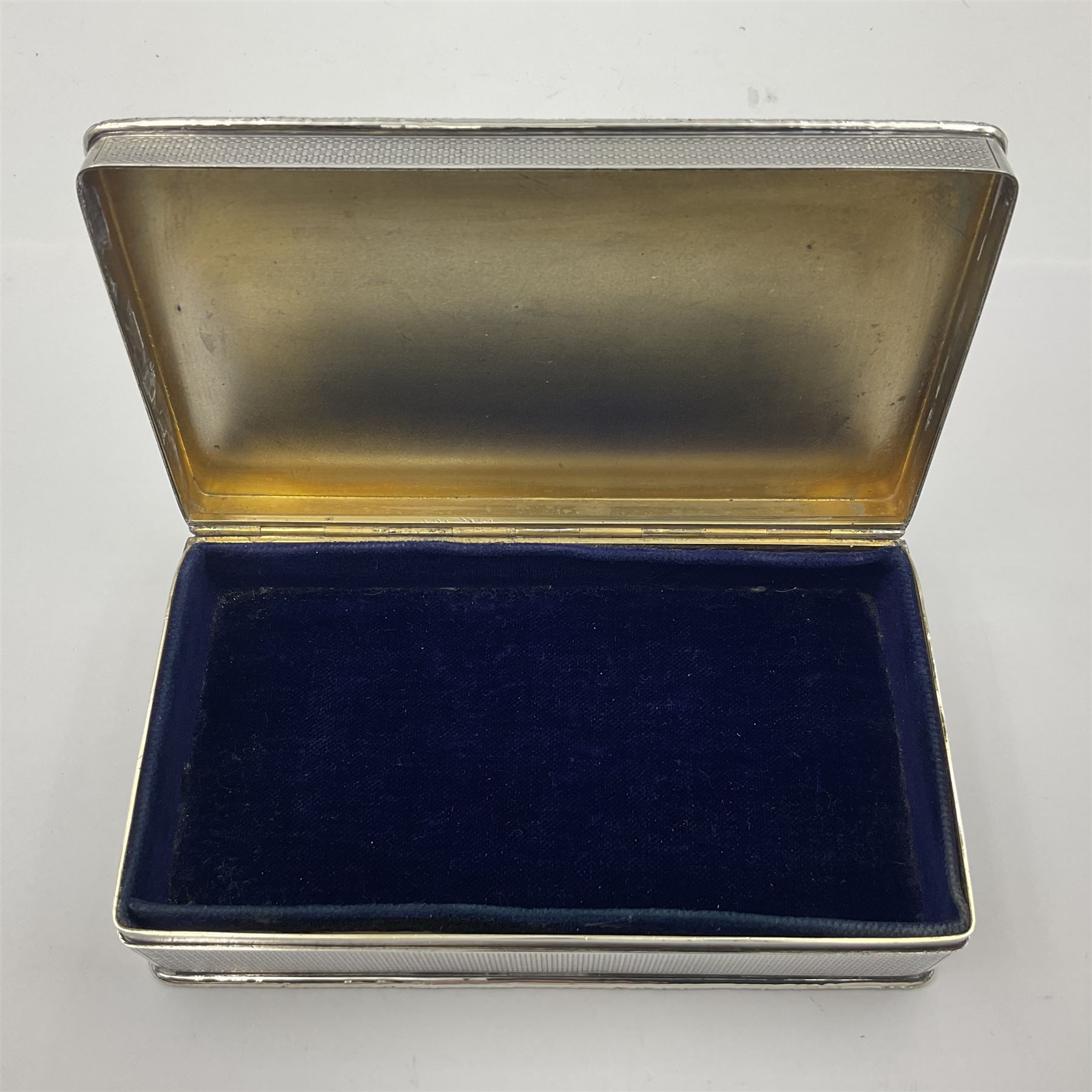 1920's silver table cigarette box, of rectangular form with engine turned decoration to the exterior, the hinged cover with central vacant rectangular panel and raised foliate rim, opening to reveal a gilt interior with later plush lining, hallmarked Charles Henry Dumenil, London 1923, H3.5cm L14.5cm D8.5cm