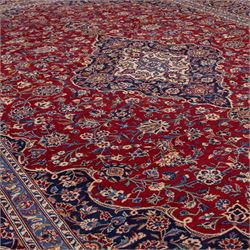 Central Persian Kashan hand-knotted carpet, madder red field with central cusped medallion in navy and ivory enclosing a floral rosette and radiating scrolling arabesques and palmettes, across a densely ornamented ground of finely drawn vinery enriched with a profusion of flowering sprays and stylized blossoms, spandrels in deep indigo and principal border of navy ground decorated with a continuous palmette and vine-scroll flanked by multiple guard stripes, with cotton fringe ends