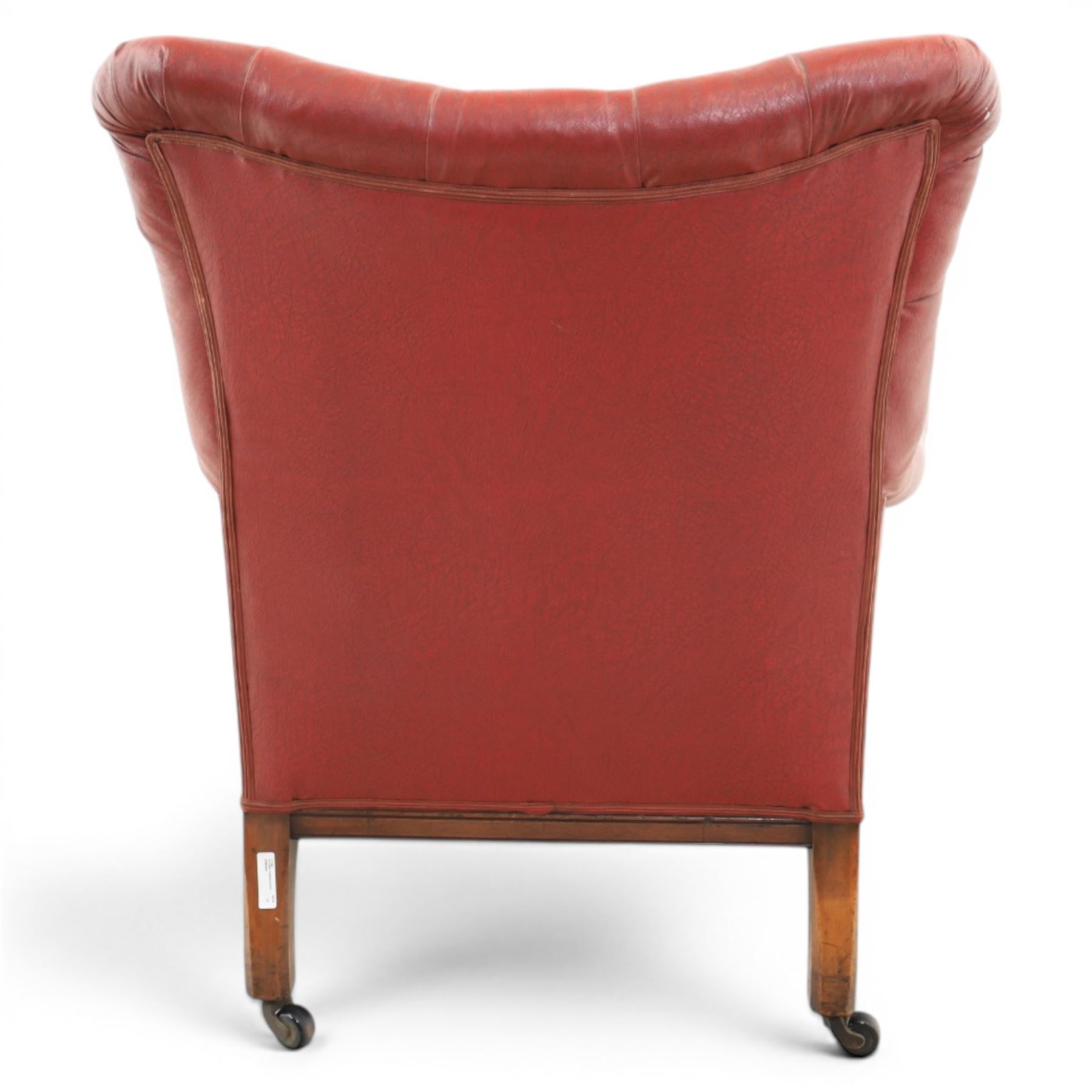Early 20th century style oxblood red leather Chesterfield armchair, deep buttoned shaped back over padded scroll arms and loose seat cushion, raised on turned mahogany front supports with brass caps and castors