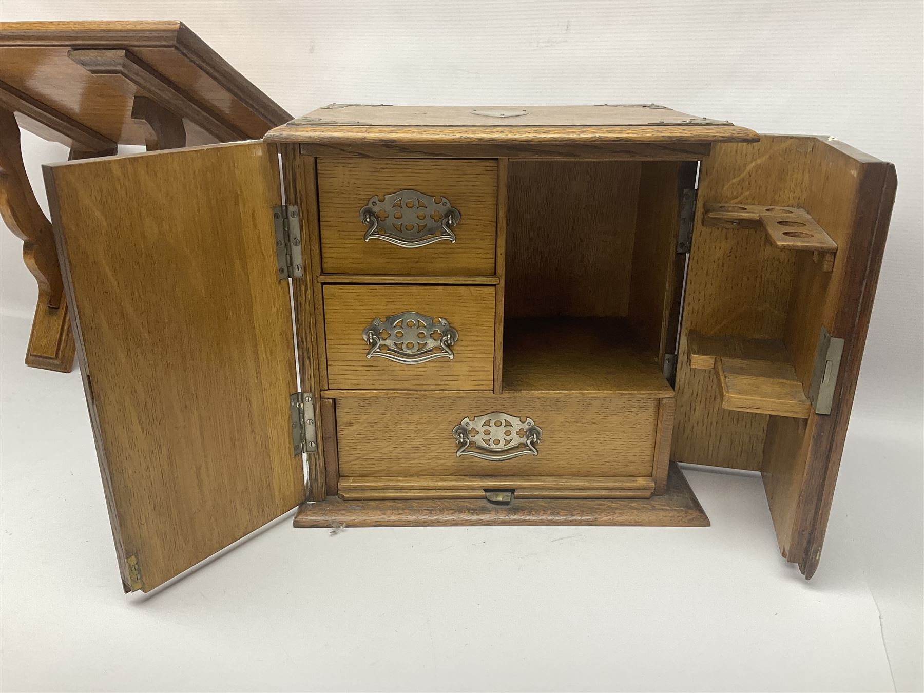 Early 20th century oak smokers cabinet, with brass shield shaped mounts, opening to reveal a compartmentalised interior with three drawers, pipe rack and tobacco jar, together with a small wooden shelf, smokers cabinet H28.5cm