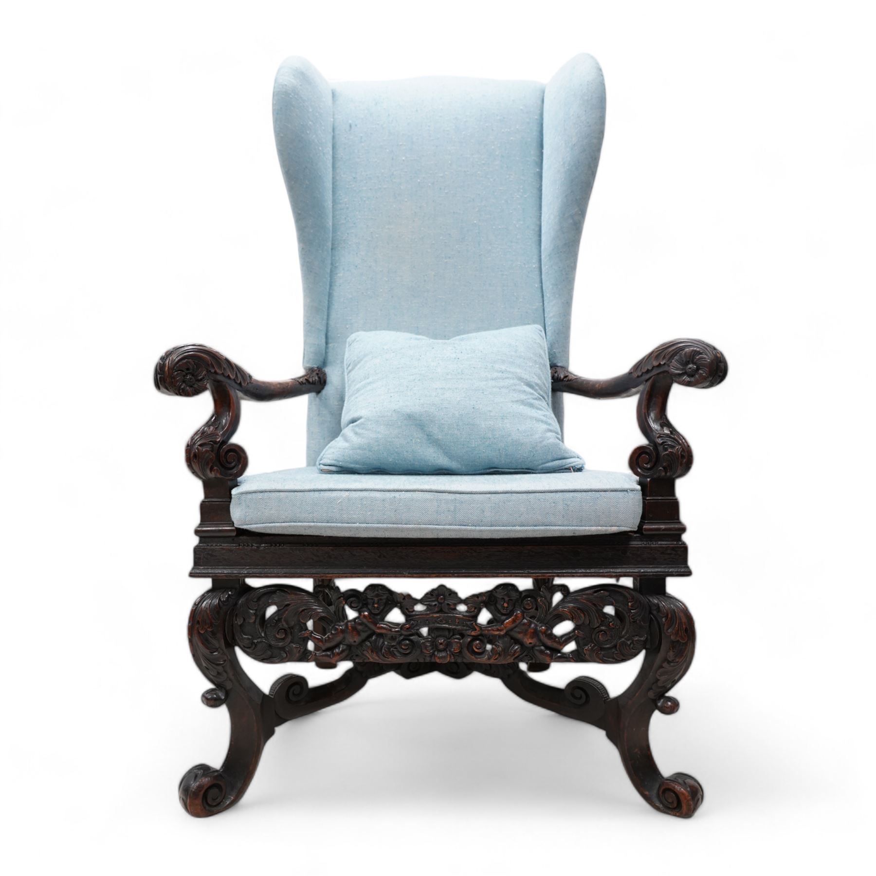 Carolean design walnut and oak wingback open armchair, the high wingback upholstered in light blue herringbone fabric, acanthus leaf carved arms with scrolled terminals and on scrolled supports, cane work seat with squab cushion and scatter cushion, the lower rail carved and pierced with putti holding crown surrounded by curled acanthus leaves, leaf carved S-scroll supports united by Scrolled X-framed stretchers 