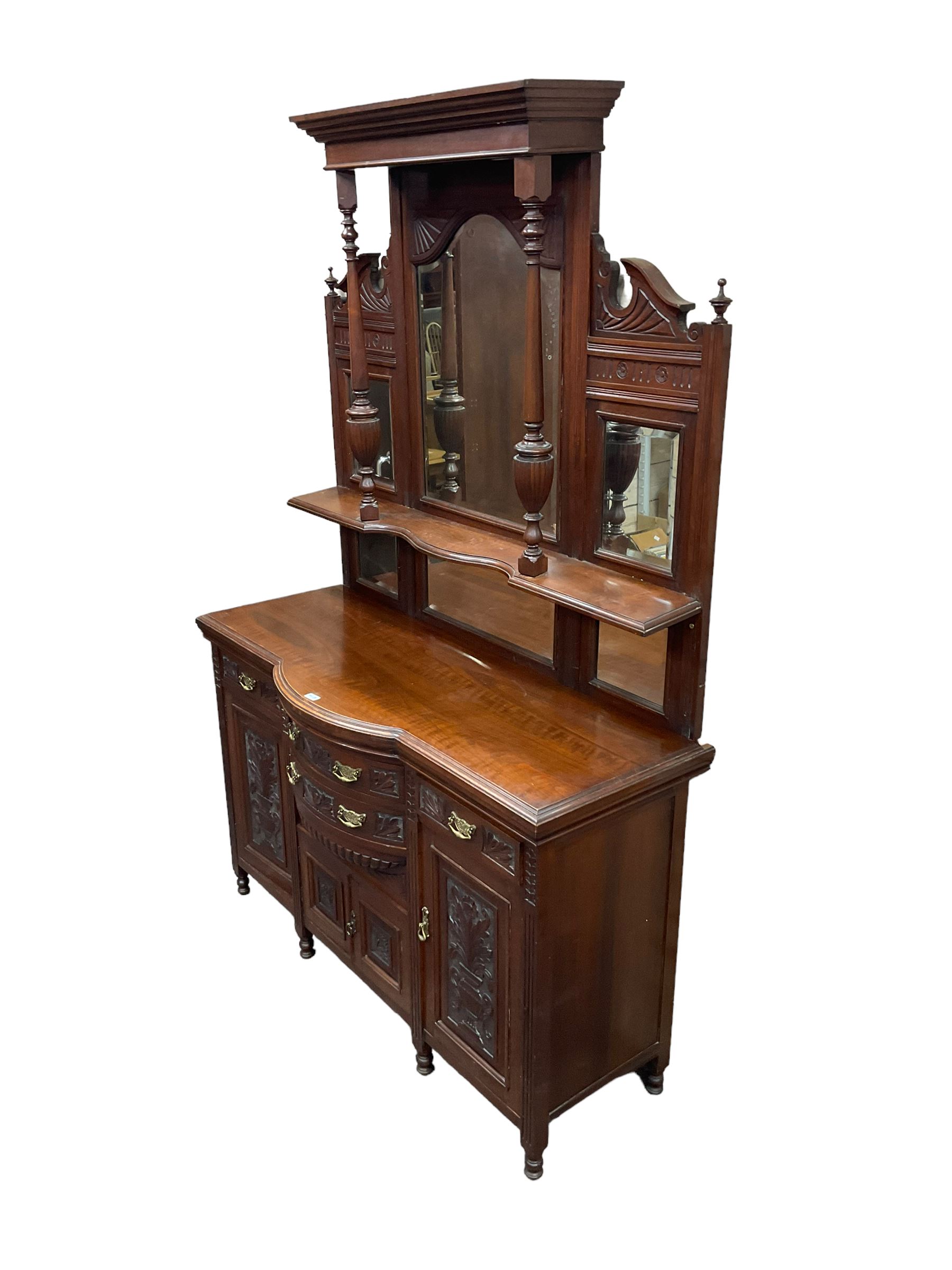 Edwardian walnut sideboard, projecting cornice supported by turned supports, the back fitted with three bevelled mirrors beneath fluted and flowerhead carved friezes, the lower section with four drawers and four panelled cupboard doors carved with foliate and urn designs, fluted uprights on turned feet