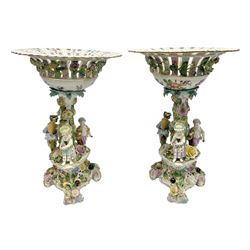 Pair of late 19th/early 20th century Continental table centre pieces, each modelled with a floral encrusted column flanked by three male and female figures with baskets of flowers, supporting a part pierced and floral encrusted bowl with hand painted floral sprays to the interior, the whole upon a conforming pierced and encrusted base with three scroll feet, each with indistinct blue mark beneath, H46cm bowl D31.5cm 