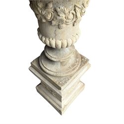 Pair of weathered composite stone campana urns on pedestals, the flared egg-and-dart rim over a tapering body with fruiting grapevine decoration in relief, over gadrooned underbelly, the rectangular pedestals moulded with floral garlands, planted