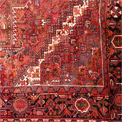 North West Persian Heriz hand-knotted carpet, madder red field, with a large central angular star medallion with stepped outlines on ivory ground, surrounded by geometric floral devices and stylized rosettes, with corresponding corner spandrels, enclosed by a bold geometric main border with multiple guard stripes, with cotton fringe ends 