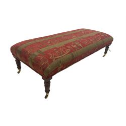 Large rectangular footstool, upholstered red and gold fabric decorated with Egyptian motifs, on turned supports with brass cups and castors; together with two matching scatter cushions 