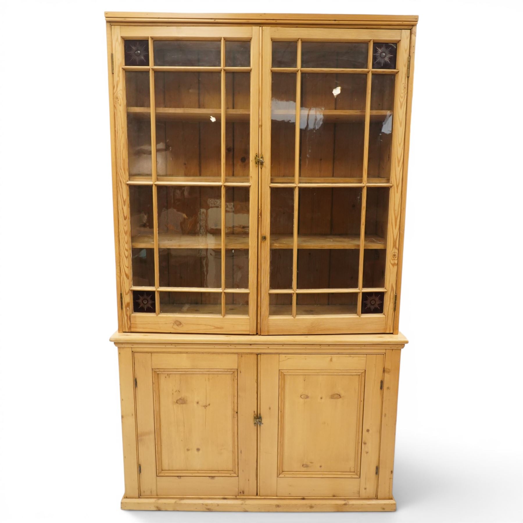 19th century pine glazed bookcase cupboard, twin astragal glazed doors with stained glass corner panels, enclosing fixed shelves, the lower section fitted with two panelled cupboard doors enclosing single shelf, on plinth base