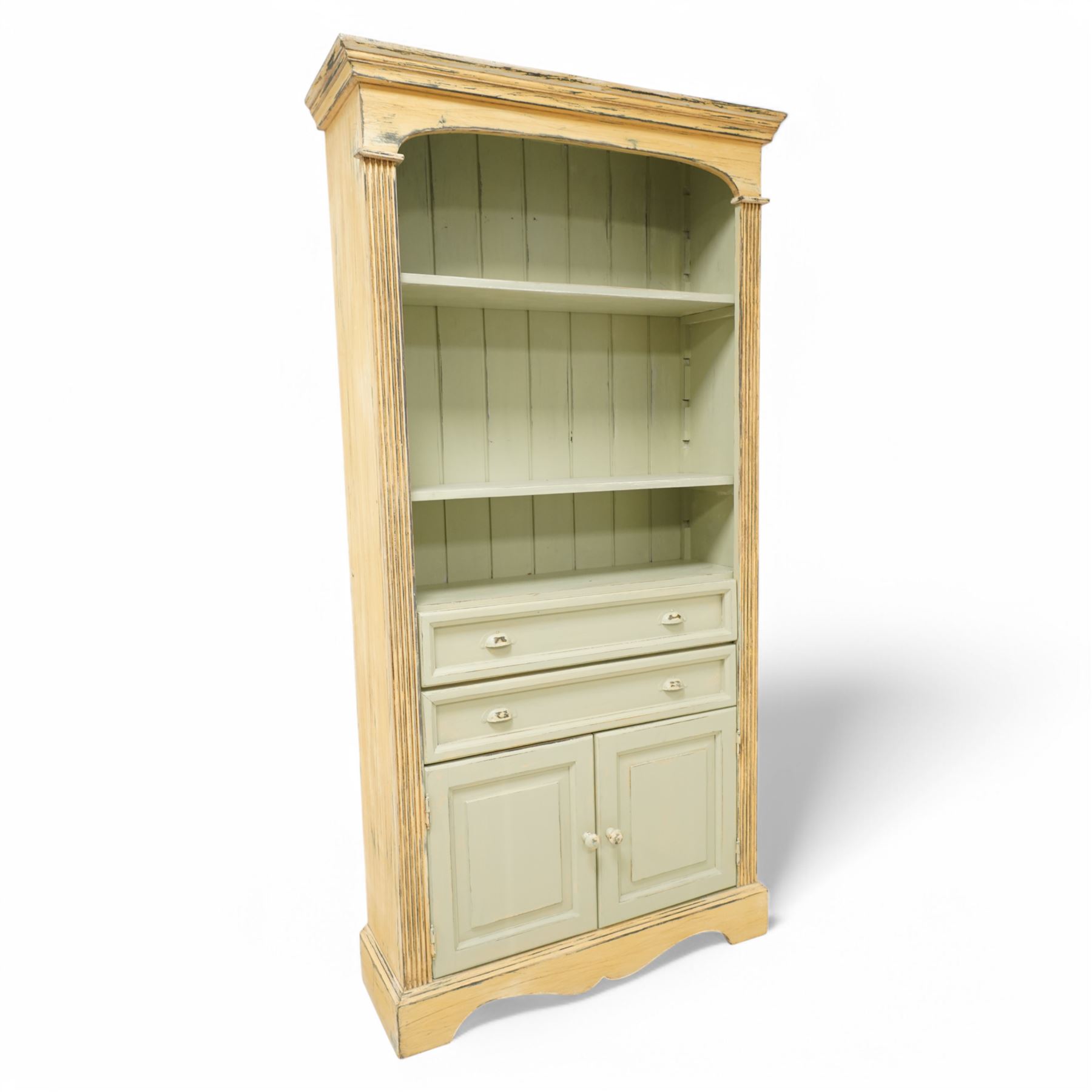Painted open bookcase over two long drawers and cupboard, with arched cornice above two shelves, in a distressed yellow and sage green 