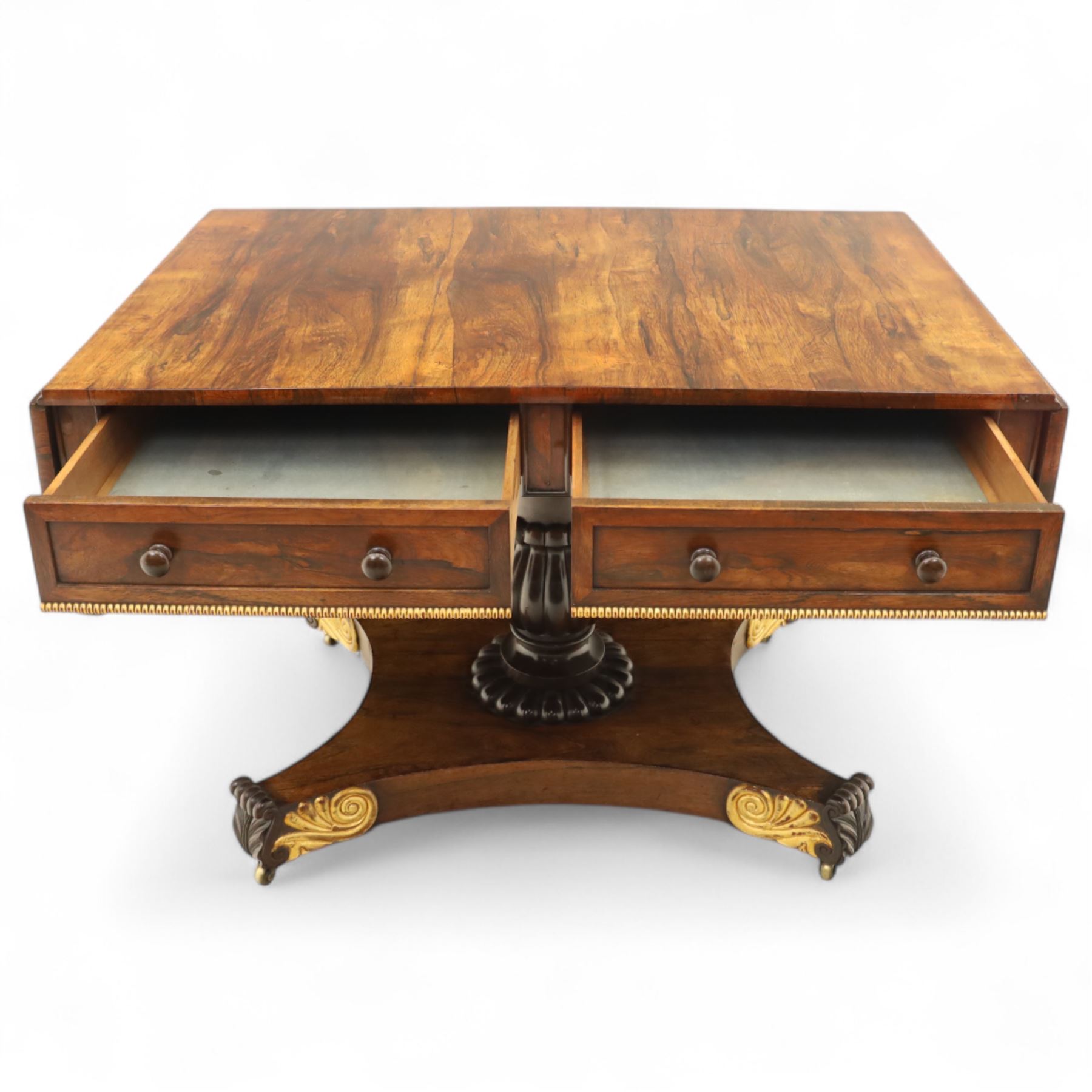 Regency rosewood sofa table, rectangular top with variegated rosewood veneers, hinged D-shaped leaves, over a set of dummy drawers to the back and full-size drawers to the front, with turned knob handles and gilt beaded moulding on frieze above elegant turned, tapering, and fluted, pedestal with a well-carved collar, supported on a large, tripartite concave platform base terminating in richly carved lion paw feet with gilded acanthus leaf carving, on concealed brass castors, circa 1815, length extended 161cm 
