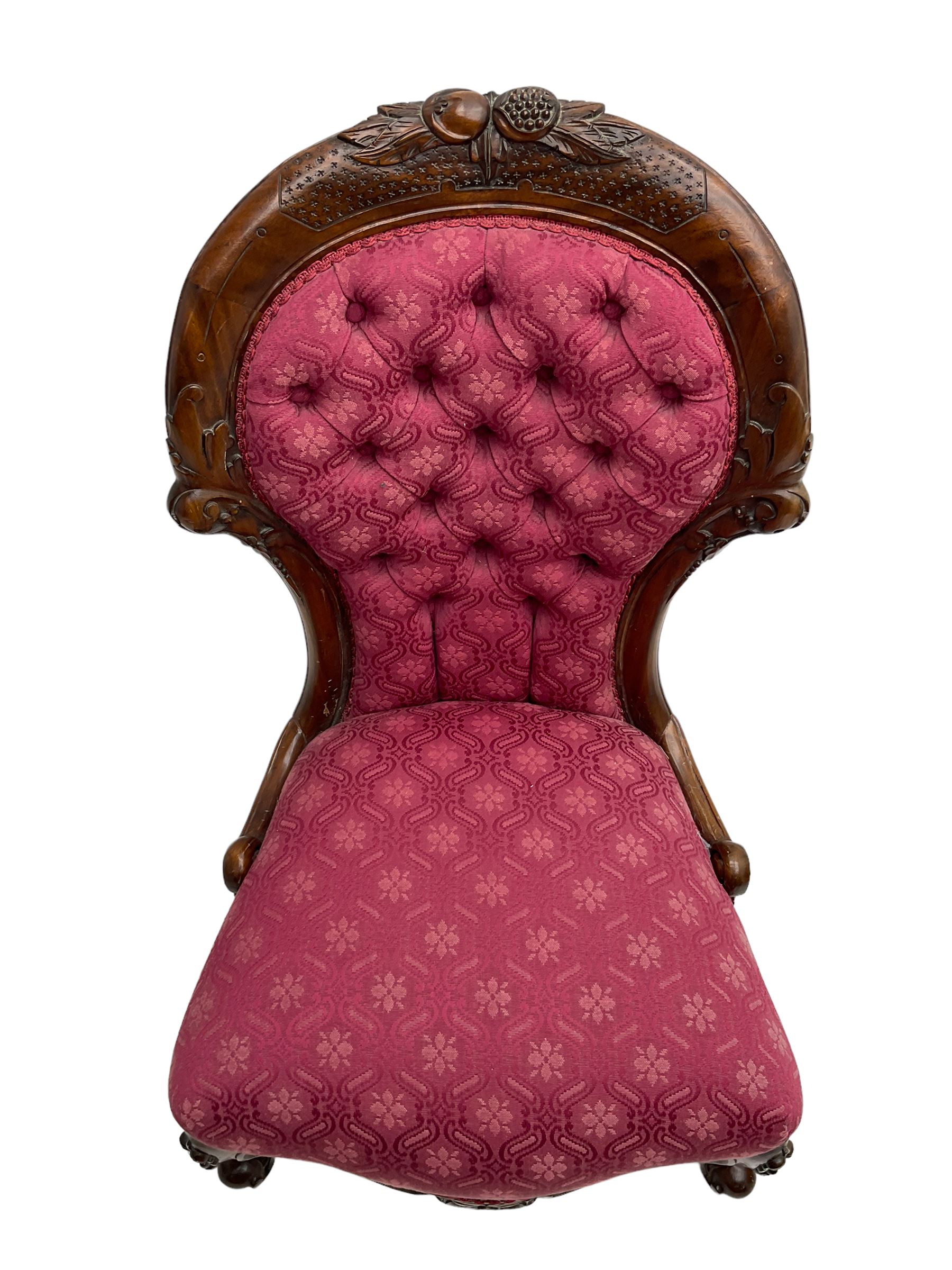 Victorian style mahogany framed nursing chair, the cresting rail carved with fruit and foliage, upholstered in red patterned fabric with buttoned back, on cabriole supports with scrolled terminals