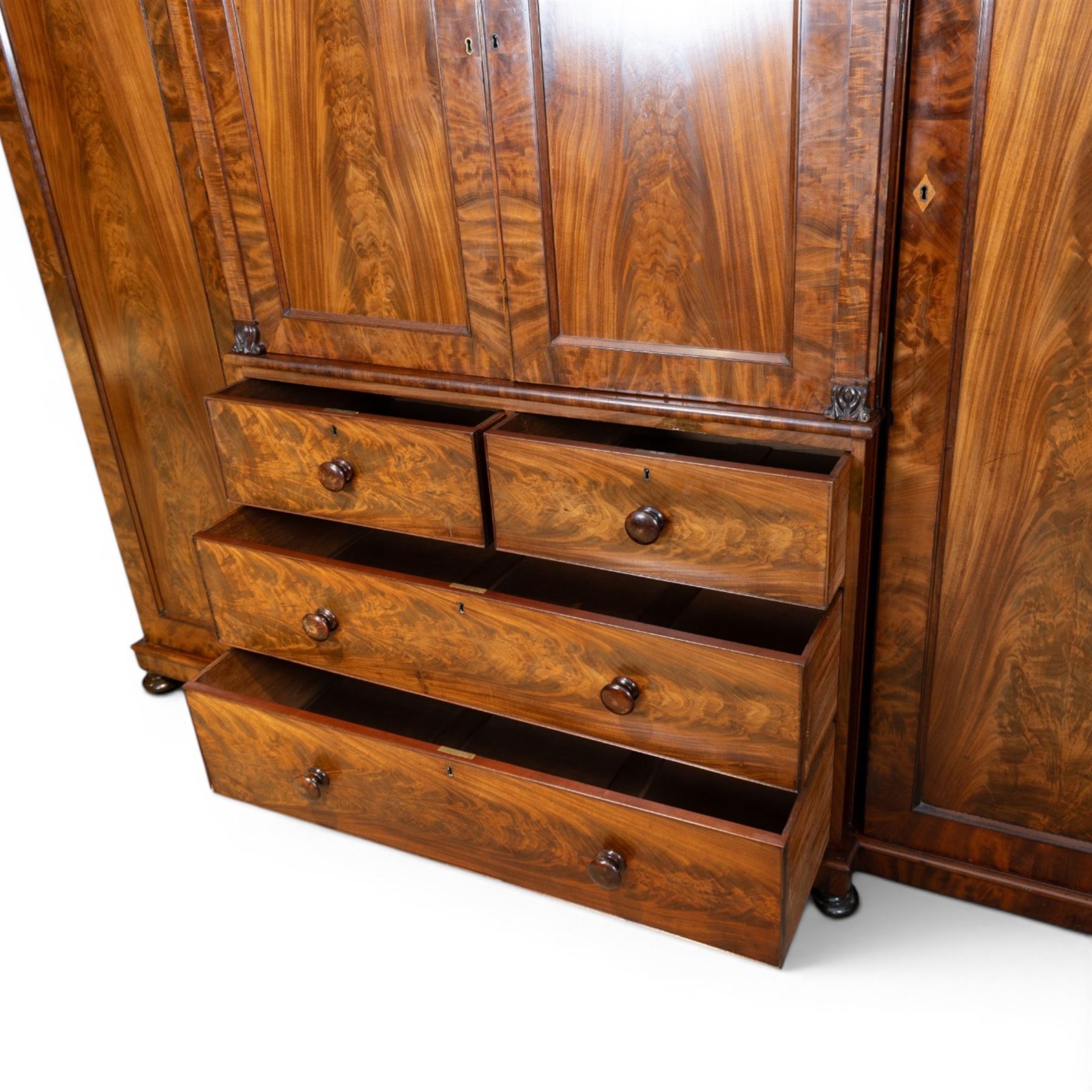 Early 19th century figured mahogany combination wardrobe, the breakfront architectural cornice over central press cupboard enclosing linen slides, flanked by full-height hanging cupboards with arched inset panels, lower arrangement of two short and two long drawers with turned knob handles, applied acanthus carved detail, raised on bun feet