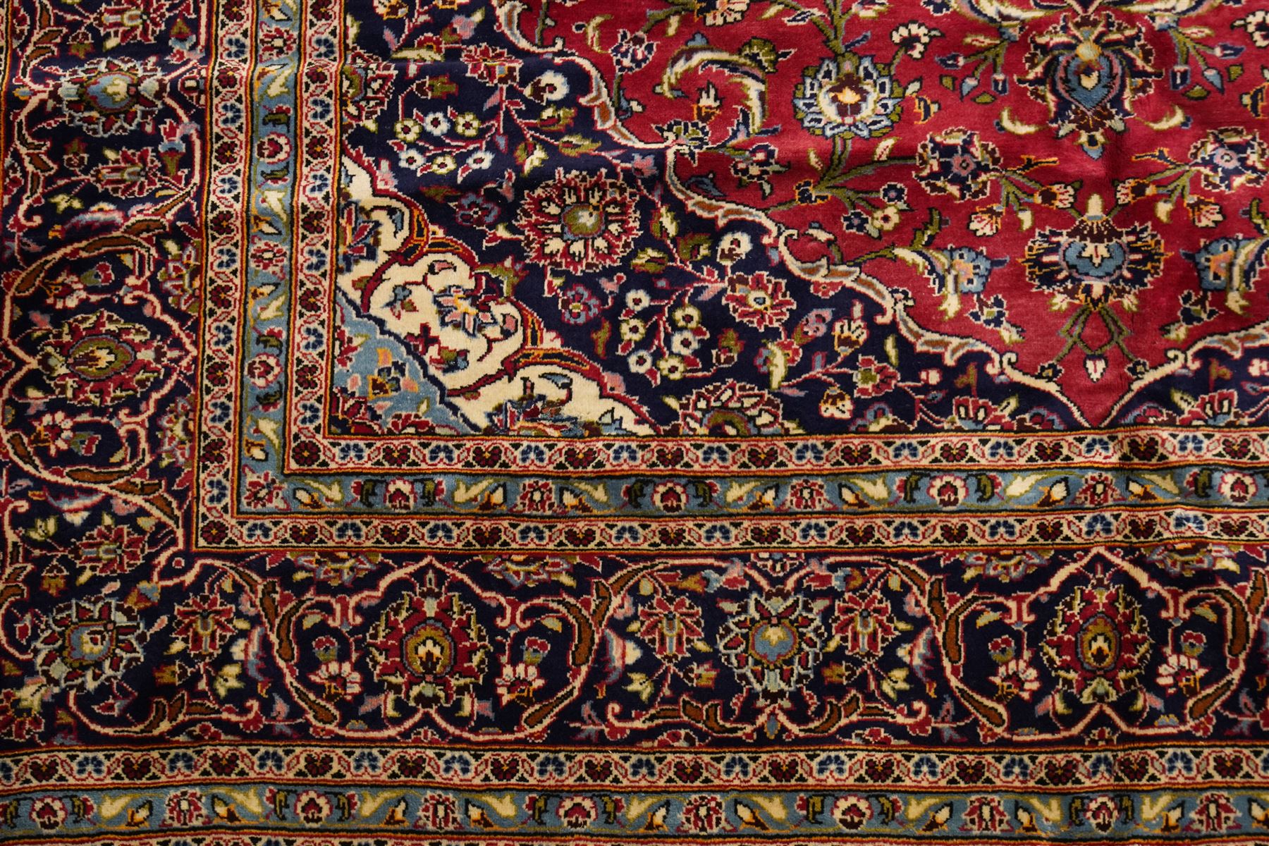 Persian Kashan hand-knotted carpet, madder red ground centred by shaped dark navy and ivory floral medallion with pendants, surrounded by scrolling vines, palmettes and flowerheads, dark navy spandrels, enclosed by dark navy floral border with multiple narrow floral guard stripes