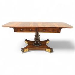 Regency rosewood sofa table, rectangular top with variegated rosewood veneers, hinged D-shaped leaves, over a set of dummy drawers to the back and full-size drawers to the front, with turned knob handles and gilt beaded moulding on frieze above elegant turned, tapering, and fluted, pedestal with a well-carved collar, supported on a large, tripartite concave platform base terminating in richly carved lion paw feet with gilded acanthus leaf carving, on concealed brass castors, circa 1815, length extended 161cm 
