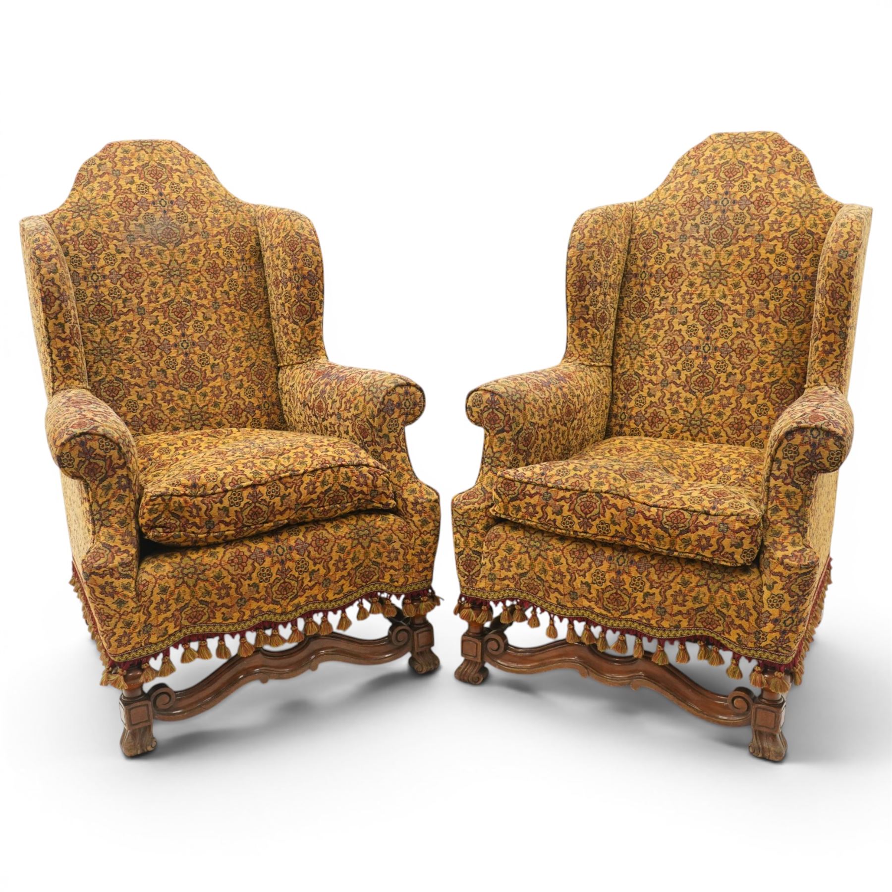 Pair of William & Mary design wingback armchairs, the arched high-back over rolled arms with a loose seat cushion and sprung seat, upholstered in old-gold ground foliate patterned fabric, raise on scroll feet united by a front serpentine stretcher