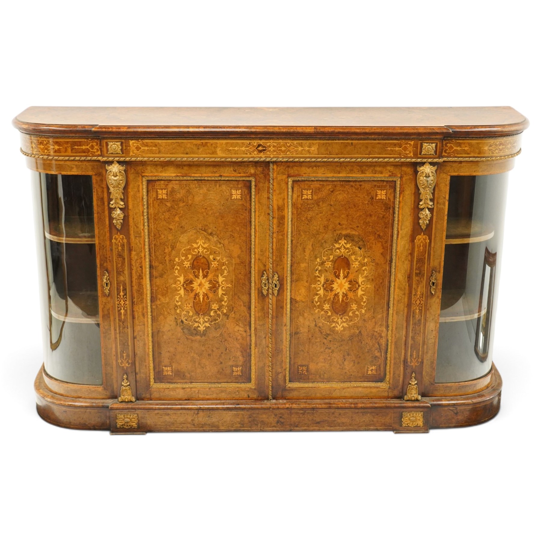 Large fine Victorian figured and burr walnut credenza, the breakfront D-shaped top with moulded edge, over an ornate frieze with inlaid Aesthetic Movement scrolling and geometric ornament, applied with gilt-metal ribbon-twist moulding and mounts, the central panelled doors profusely inlaid with scrolling foliate motifs and stringing, flanked by gilt-metal cartouche and scrolling acanthus corbel mounts, with flanking glazed bow-front side doors, on skirted base with further gilt metal foliate mounts