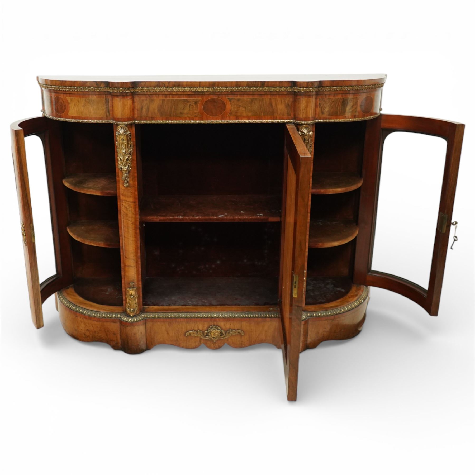 Late Victorian figured walnut credenza, the shaped demi-lune top with cross-banding and stringing over an inlaid frieze with foliate gilt-metal mounts, fitted with a central geometric-inlaid panelled door, flanked by convex-glazed side-cabinets with a gilt metal slip and outer-banding, the uprights surmounted by gilt-metal cartouche mounts with cascading foliage details, raised on a shaped skirted base with central female-mask gilt mount