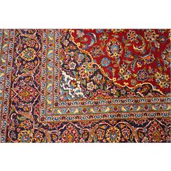 Central Persian Kashan hand-knotted carpet, crimson field with central cusped medallion in ivory, pale blue and indigo, issuing a network of scrolling arabesques, palmettes, and floral sprays, with conforming spandrels, main border of scrolling vinery and stylized palmettes, framed by multiple guard borders, with cotton fringe ends 