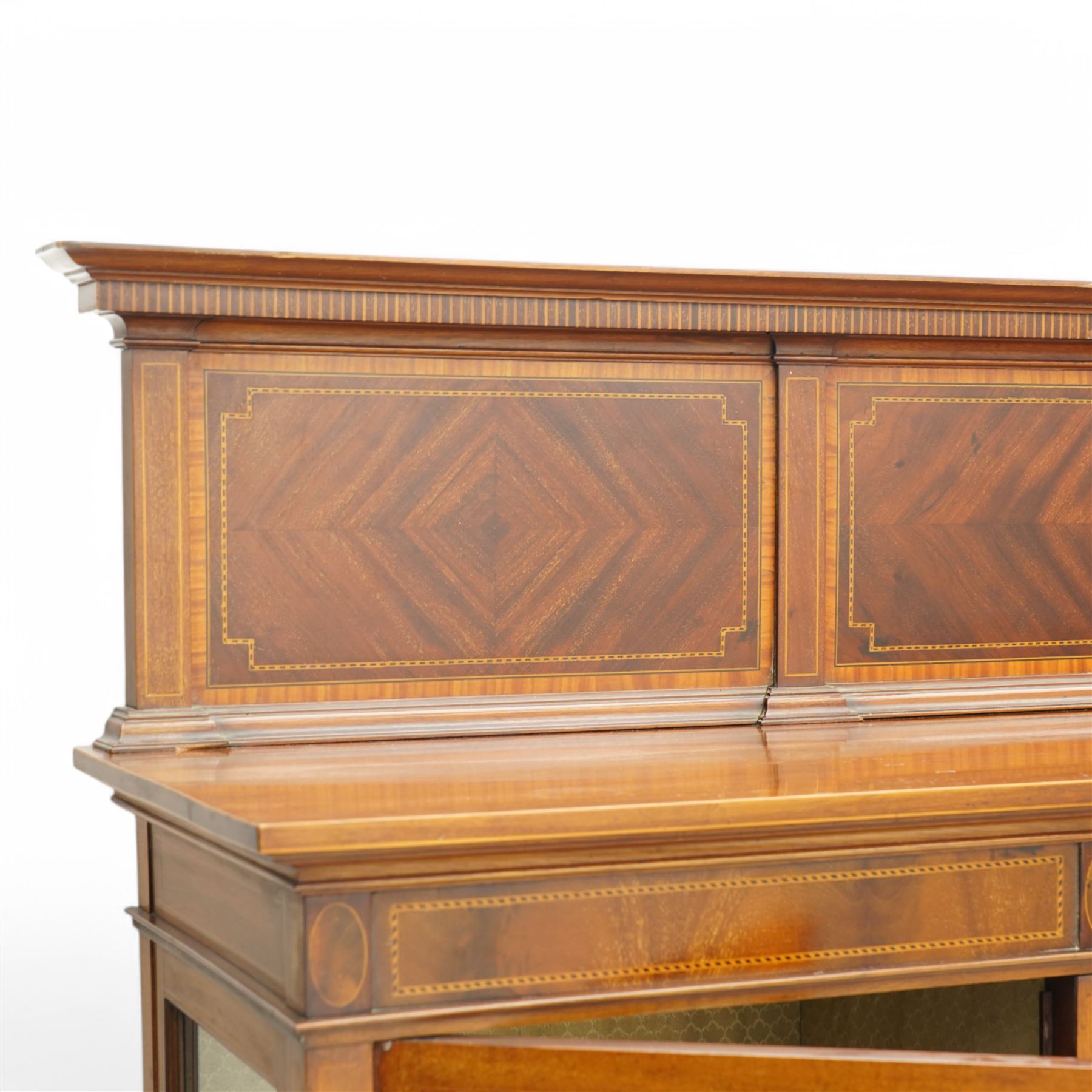Maple & Co. Edwardian inlaid mahogany display cabinet, raised triple panelled back inlaid with quarter-matched veneers, break bow-fronted top over figured frieze with checkered stringing, enclosed by two astragal glazed doors and central panelled door, oval fan inlay with ribbon tie and trailing bell flowers, on square tapering supports with spade feet united by under tier, with 'Maple & Co.' circular makers plaque to the back