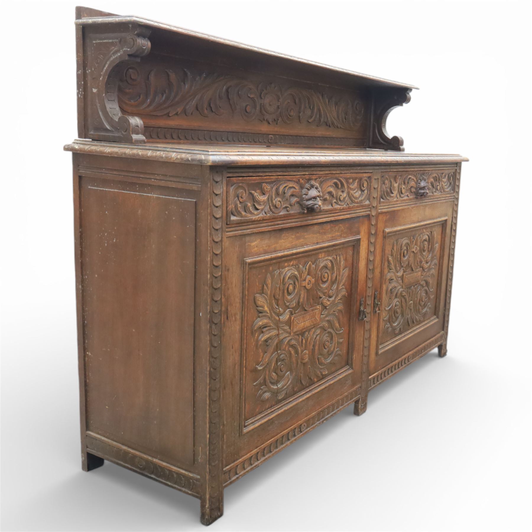 Victorian carved oak sideboard, the rectangular top with raised back carved with foliate scrolls and scroll end supports, upon a base with rectangular top with moulded edge, pair of drawers with conforming carved decoration and mask handles, and pair of carved cupboard doors, upon short stile type feet H101.5cm W183cm D54cm 