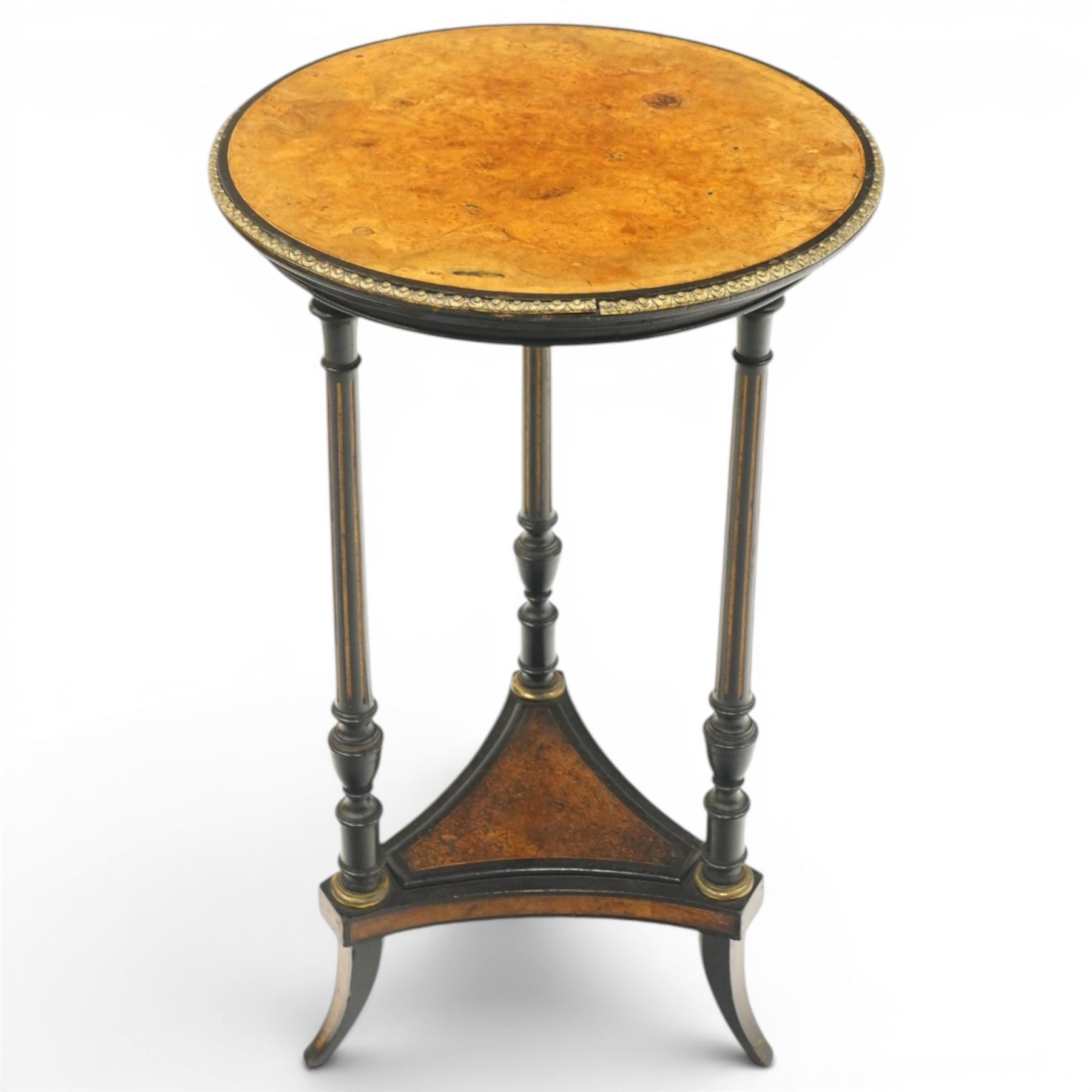 Late Victorian Aesthetic Movement ebonised and figured walnut occasional table, the circular burr top with boxwood strung and ebonised edge, surrounded by gilt metal egg-and-dart moulding, raised on three turned and fluted pedestals with parcel-gilt decoration, united by a triform undertier raised on splayed feet