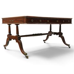 Regency inlaid mahogany and brass mounted library table, the rectangular top inset with gilt tooled burgundy leather writing surface, enclosed by  crossbanding and boxwood stringing with roundel corner motifs, above three cock-beaded frieze drawers to either side fitted with brass ring handles and brass escutcheons, the end friezes centred by applied brass foliate mounts, raised on twin reeded column supports over rectangular platform ends, united by a ring-turned stretcher, the reeded sabre legs terminating in brass caps and castors, one drawer bearing ivorine label 'Spillman & Co., St Martins Lane, London'