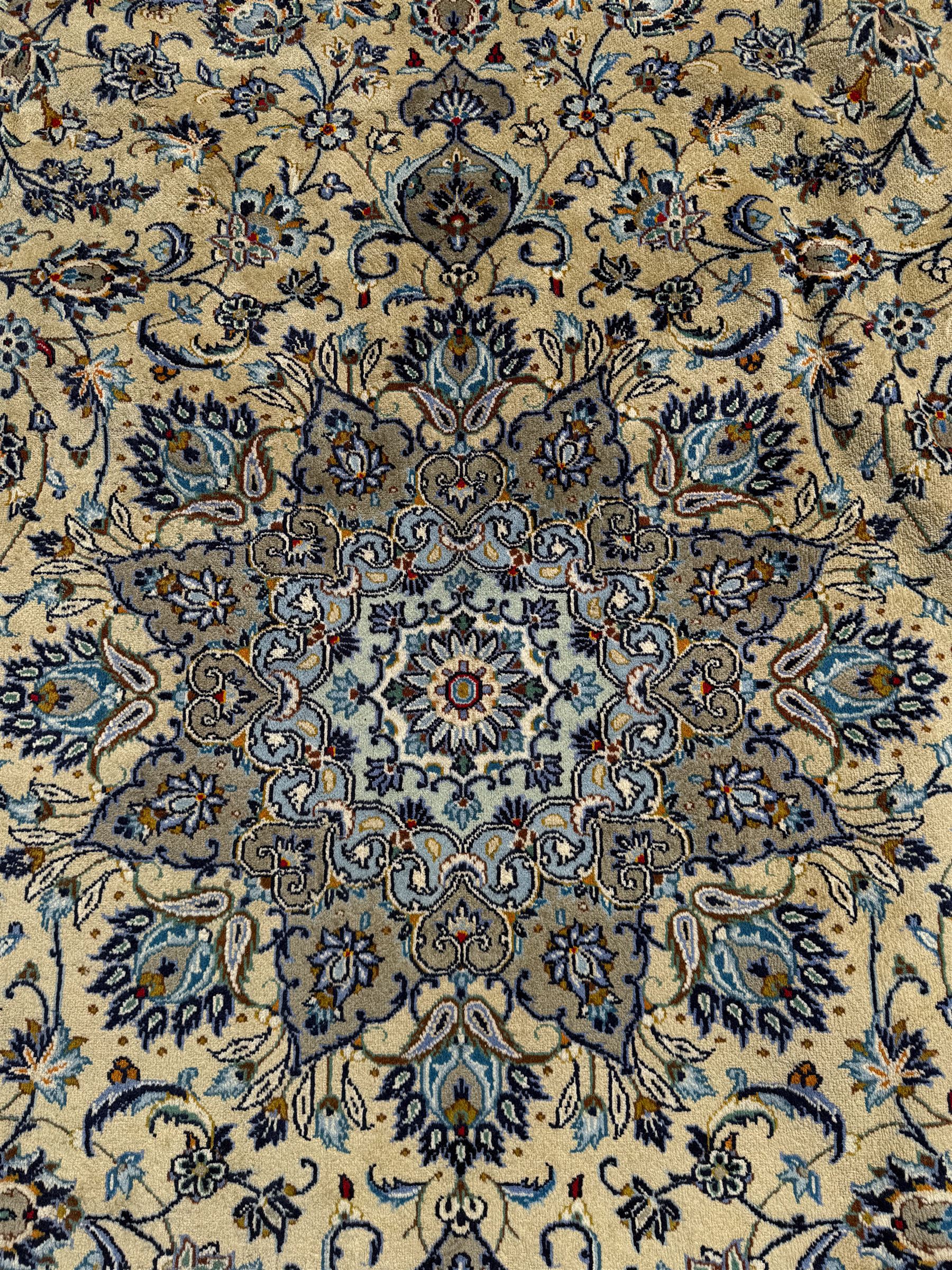 Central Persian Kashan pale khaki and ivory ground carpet, shaped central medallion enclosing small stylised motifs, the field decorated profusely with trailing foliate branches and plant motifs, guarded border with repeating floral design