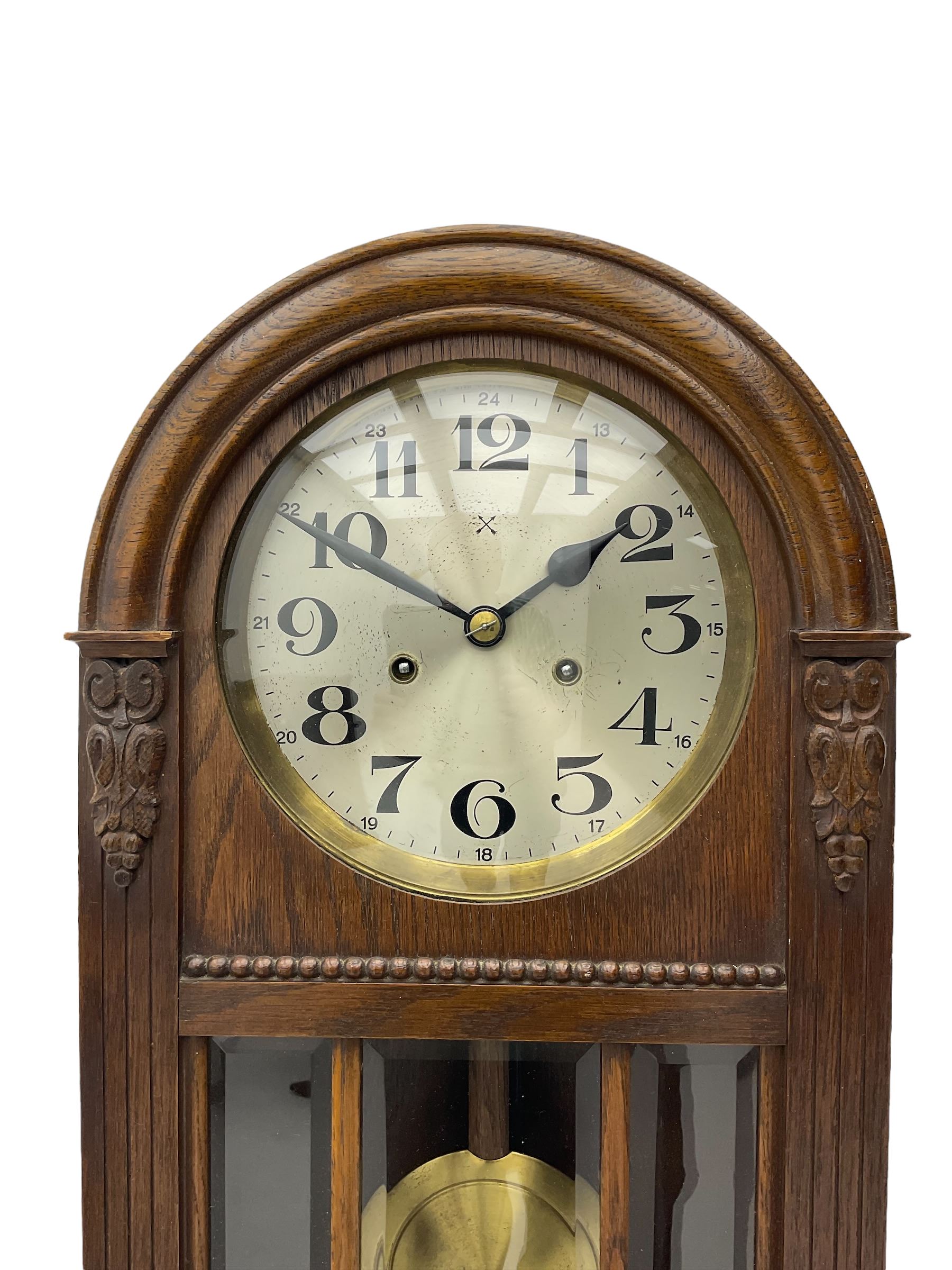 German HAC “ting tang” quarter striking wall clock in a round arched oak case with applied carving and beadwork, glazed door with a visible pendulum and silvered 24-hour dial with Arabic numerals and steel spade hands,  8-day spring driven movement striking on gong rods.

