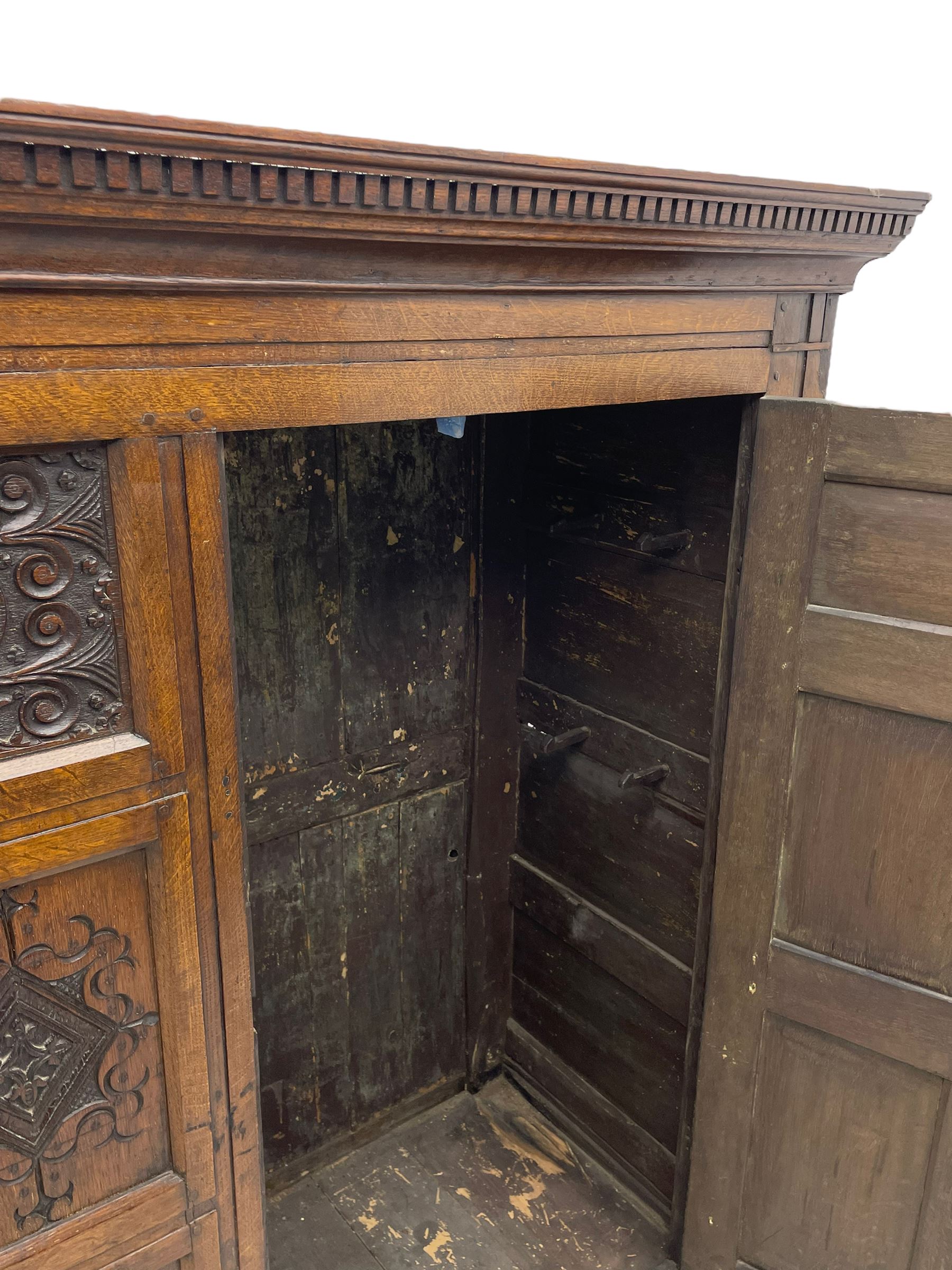 Large 18th century and later oak livery cupboard, projecting moulded and dentil cornice, sunken fillet moulded frame, triple vertical panelled central upright, enclosed by two doors each with five panels, the top row of panels carved with stylised scrolls, the lower panels carved with foliate lozenges, on shaped bracketed skirt base 