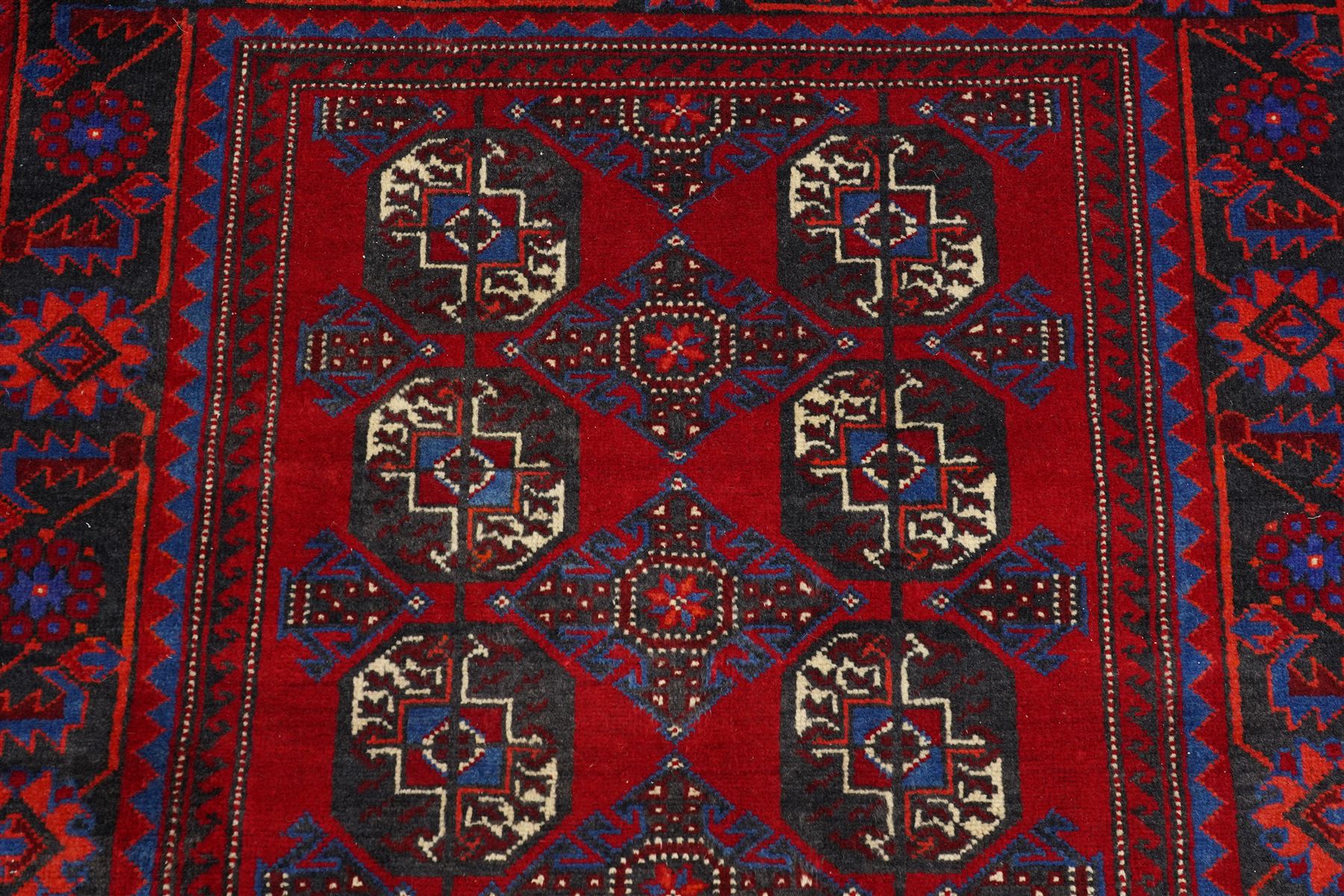 North East Persian Balochi hand-knotted runner, crimson field decorated with three vertical columns of repeating octagonal guls, each enclosing angular geometric motifs in deep indigo, ivory, and soft brown tones, guls linked by stepped diamond and cruciform devices, main border of stylized geometric floral motifs and angular vines, flanked by multiple guard borders, with cotton fringe ends 