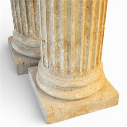 A pair of early 20th century Italian Neoclassical pedestal columns of simplified Doric type; each of fluted cylindrical form in composite stone, finished to evoke weathered limestone or travertine, raised on square plinth bases