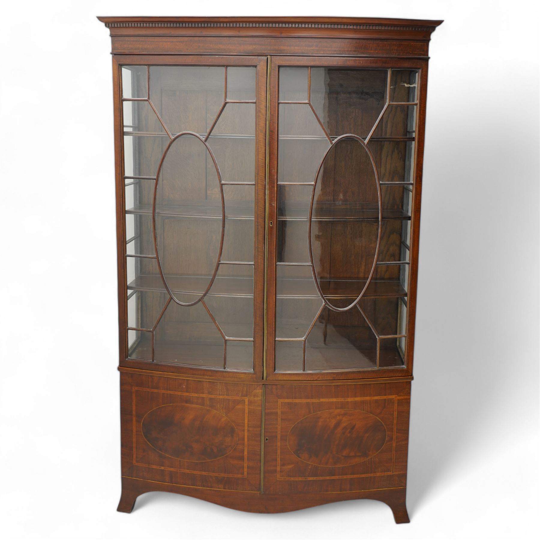 Edwardian mahogany and satinwood bow-fronted display cabinet, moulded dentil cornice over two astragal glazed doors, fitted with three shelves, double cupboard below, the doors inlaid with figured oval panels and satinwood bands, shaped apron on splayed bracket feet 