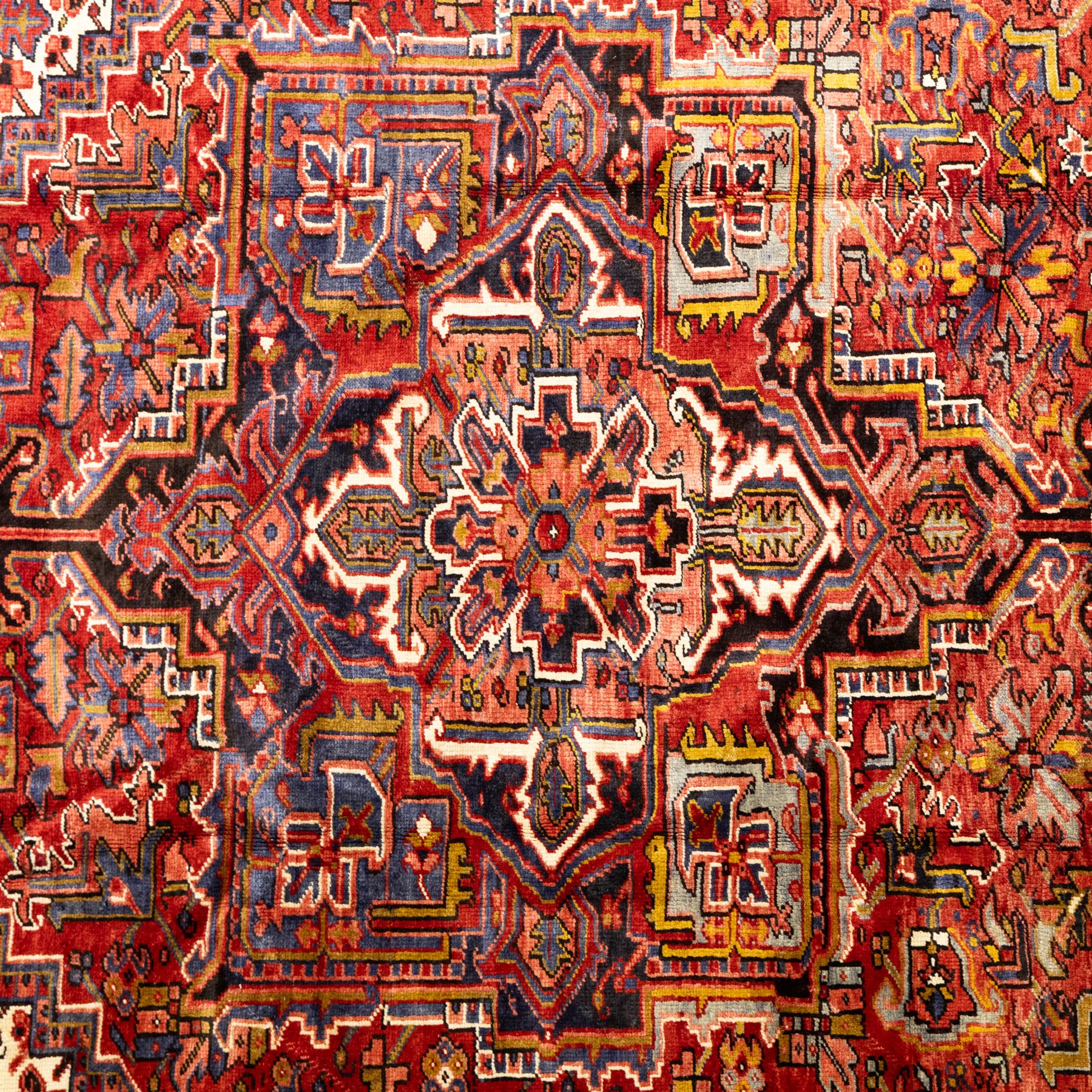 North West Persian Heriz hand-knotted carpet, madder red ground, with a large central geometric medallion with pendants, surrounded by angular foliate and floral motifs incorporating stylised rosettes, palmettes and serrated vine work, enclosed by ivory spandrels and a dark navy border with repeating geometric flowerheads and angular meandering vines, flanked by narrow patterned guard stripes, with cotton fringes